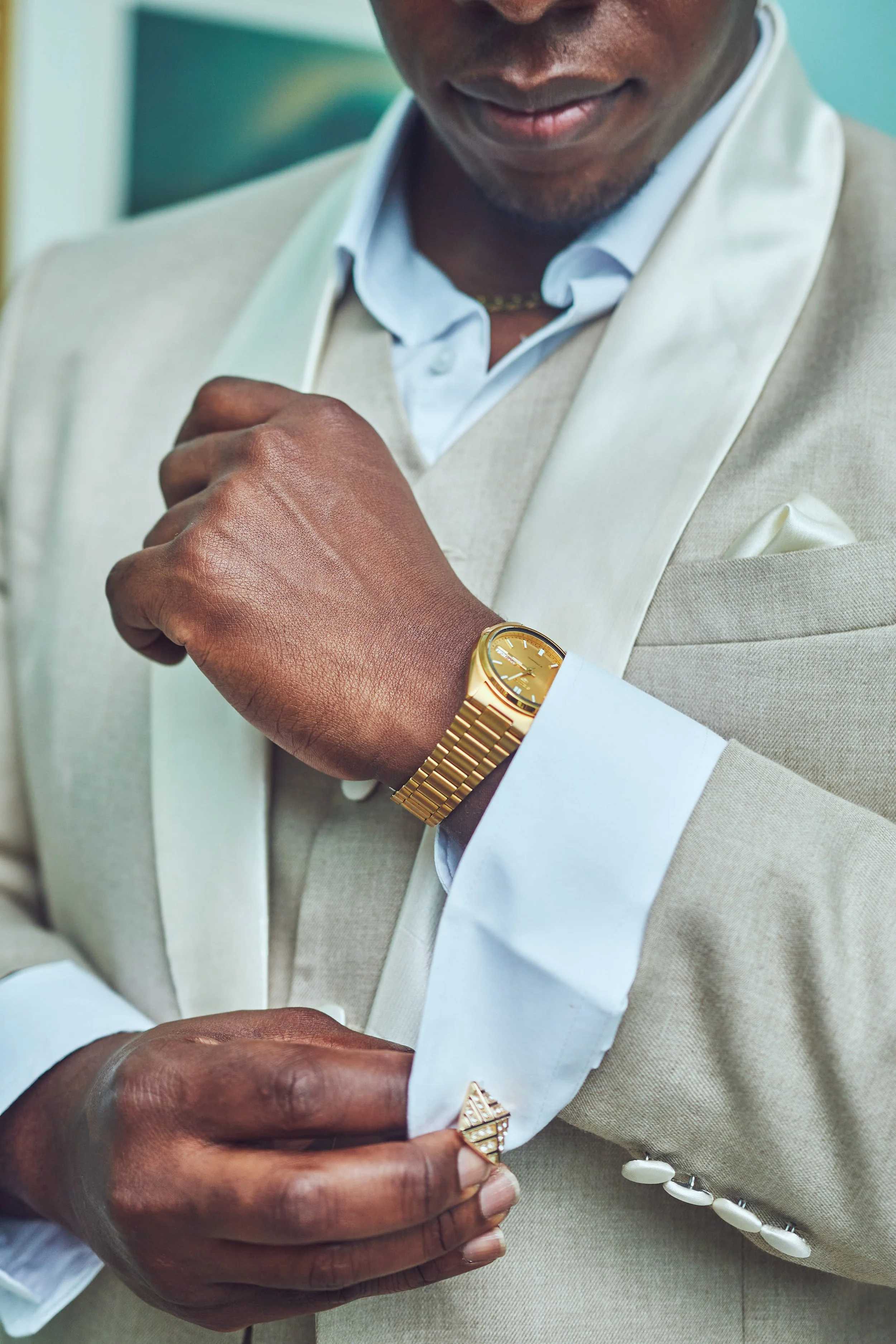 A man in a beige tuxedo adjusting his white shirt sleeve, wearing a gold wristwatch and a gold ring.