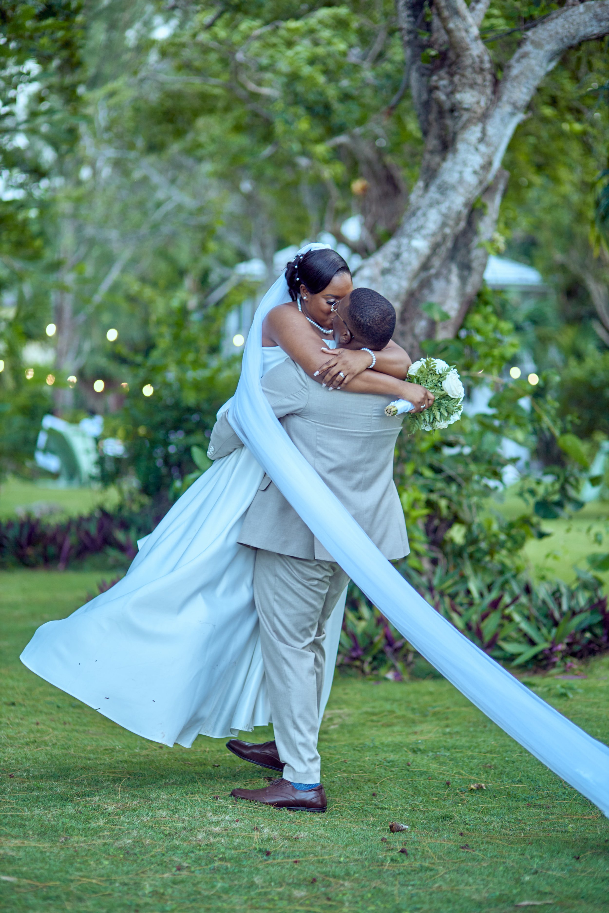 A bride and groom share a kiss outdoors, the groom lifting the bride, who is holding a bouquet of white roses, in a lush garden setting with trees and greenery.