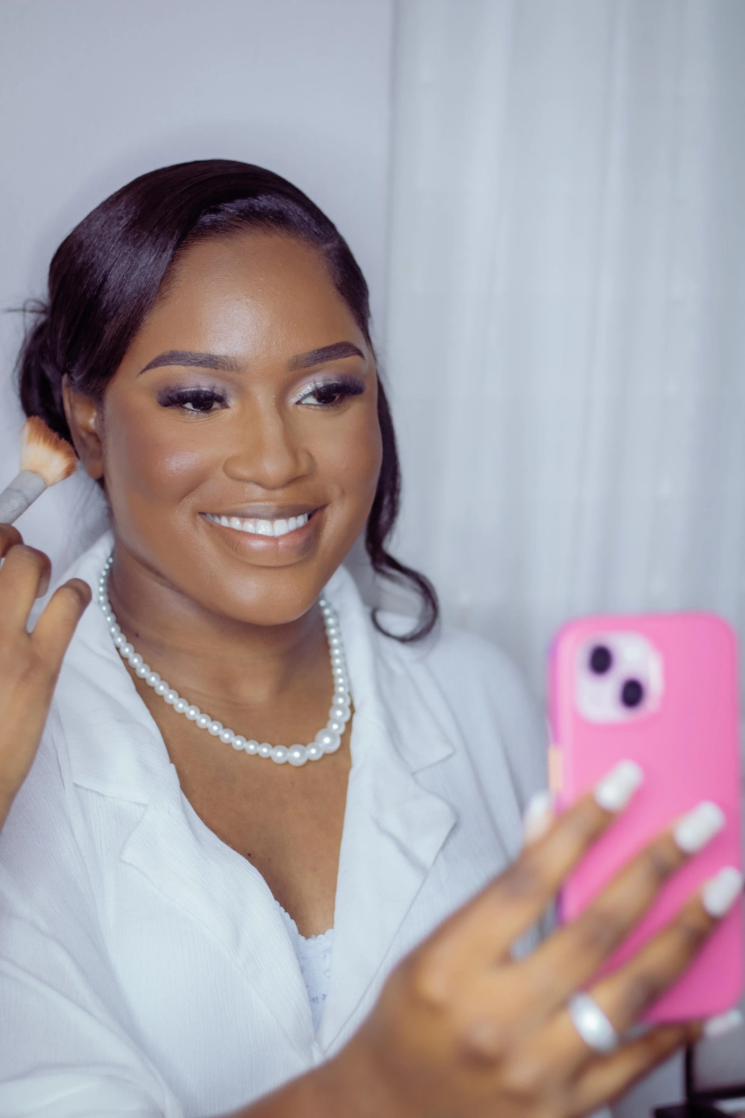 A woman with dark hair and makeup is looking at her phone while smiling, holding a makeup brush near her face.