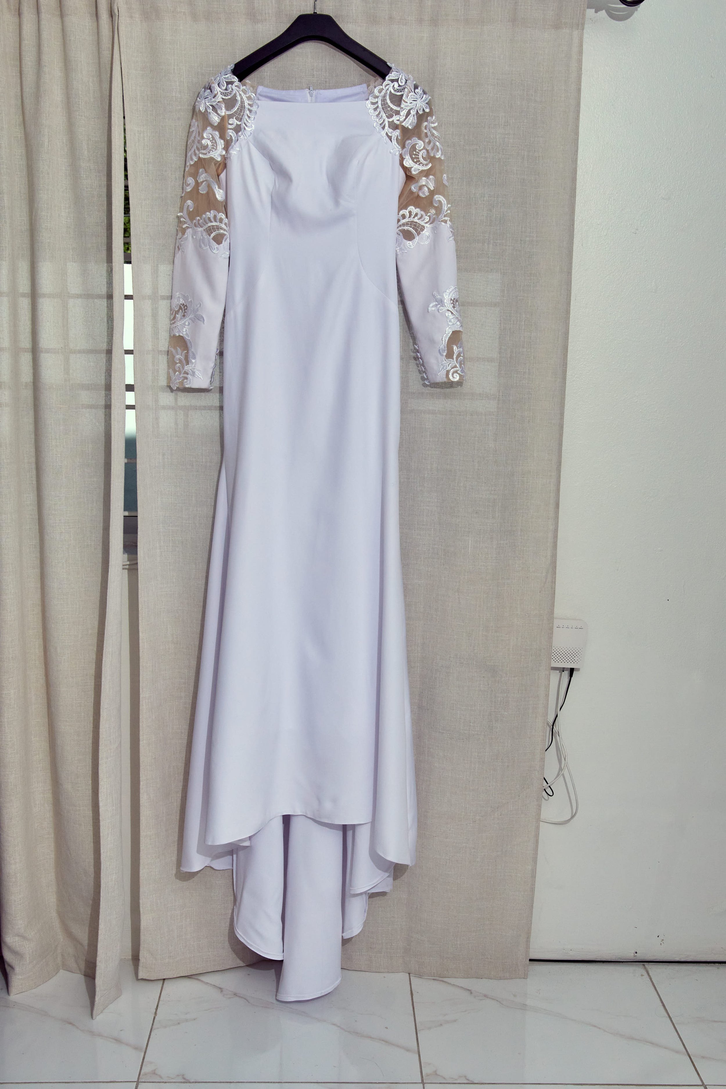 White wedding dress with lace embroidered sheer long sleeves hanging on a black hanger against beige curtains and a white wall.