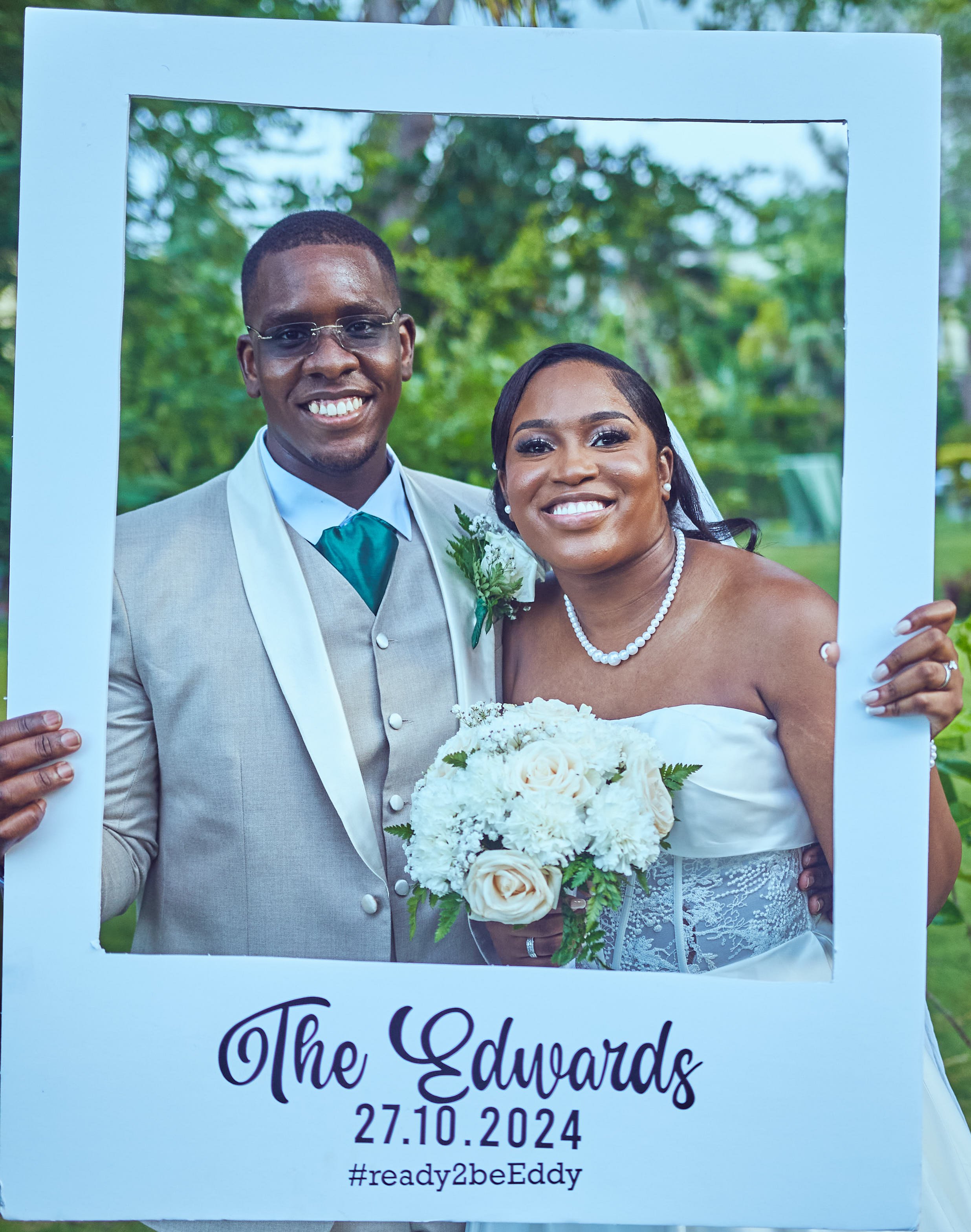 A newlywed couple holding a white frame with wedding details, smiling outdoors. The groom wears a gray suit, teal tie, and glasses, while the bride wears a strapless white wedding dress, pearl necklace, and holds a bouquet of white flowers. The frame