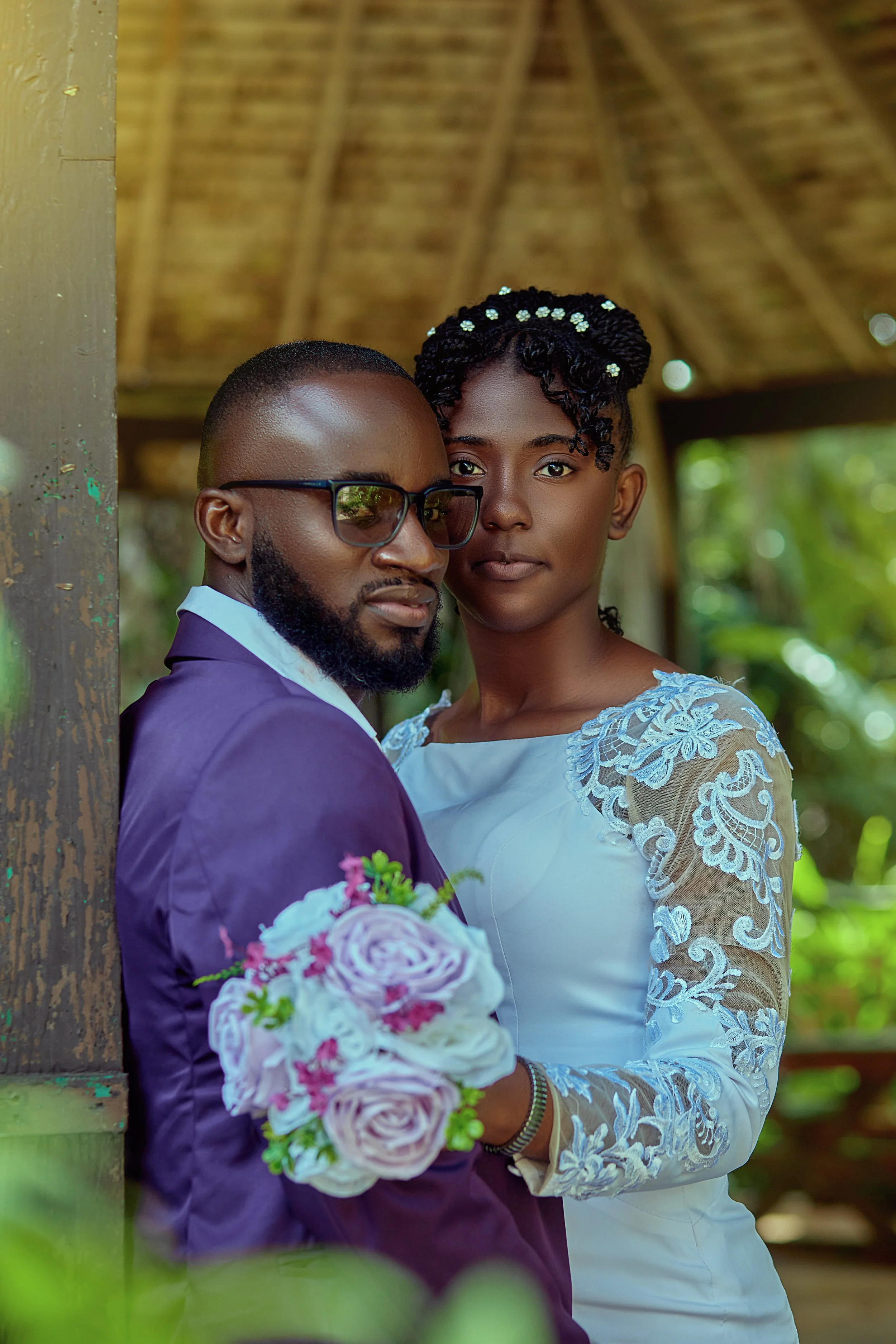 A couple dressed in formal attire, with the man wearing sunglasses and holding a bouquet of pink and white roses, standing close together in a rustic outdoor setting with a thatched roof background.
