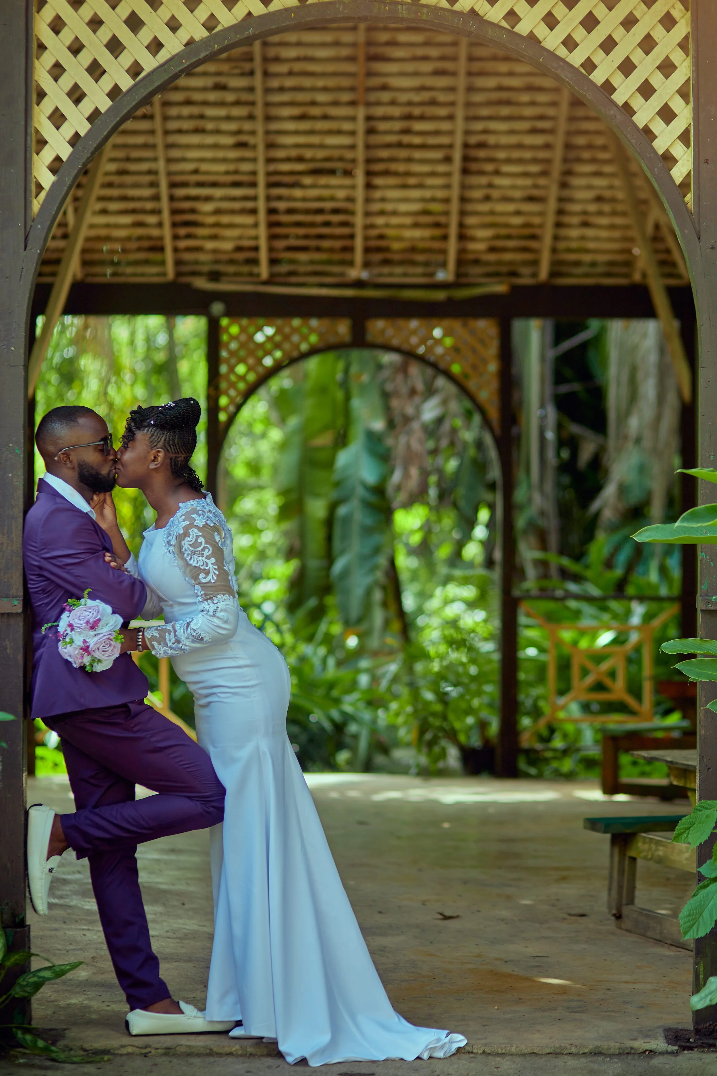 A couple in wedding attire kissing under a wooden archway in a lush, green outdoor setting.