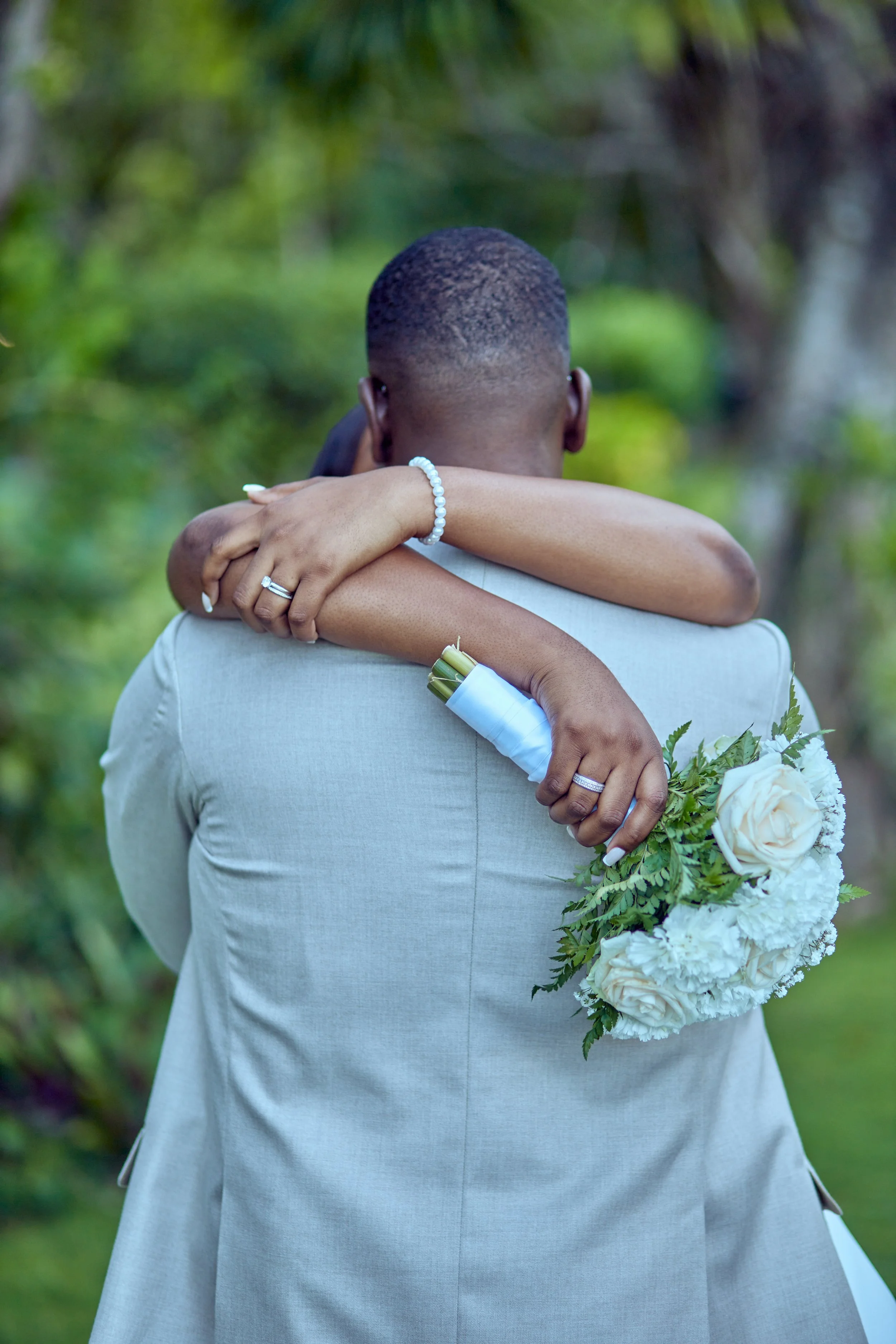 Back of a couple hugging; the woman holds a bouquet of white roses and greenery, and wears a pearl bracelet and wedding rings; the man wears a light gray suit. The background shows blurred green trees and plants.