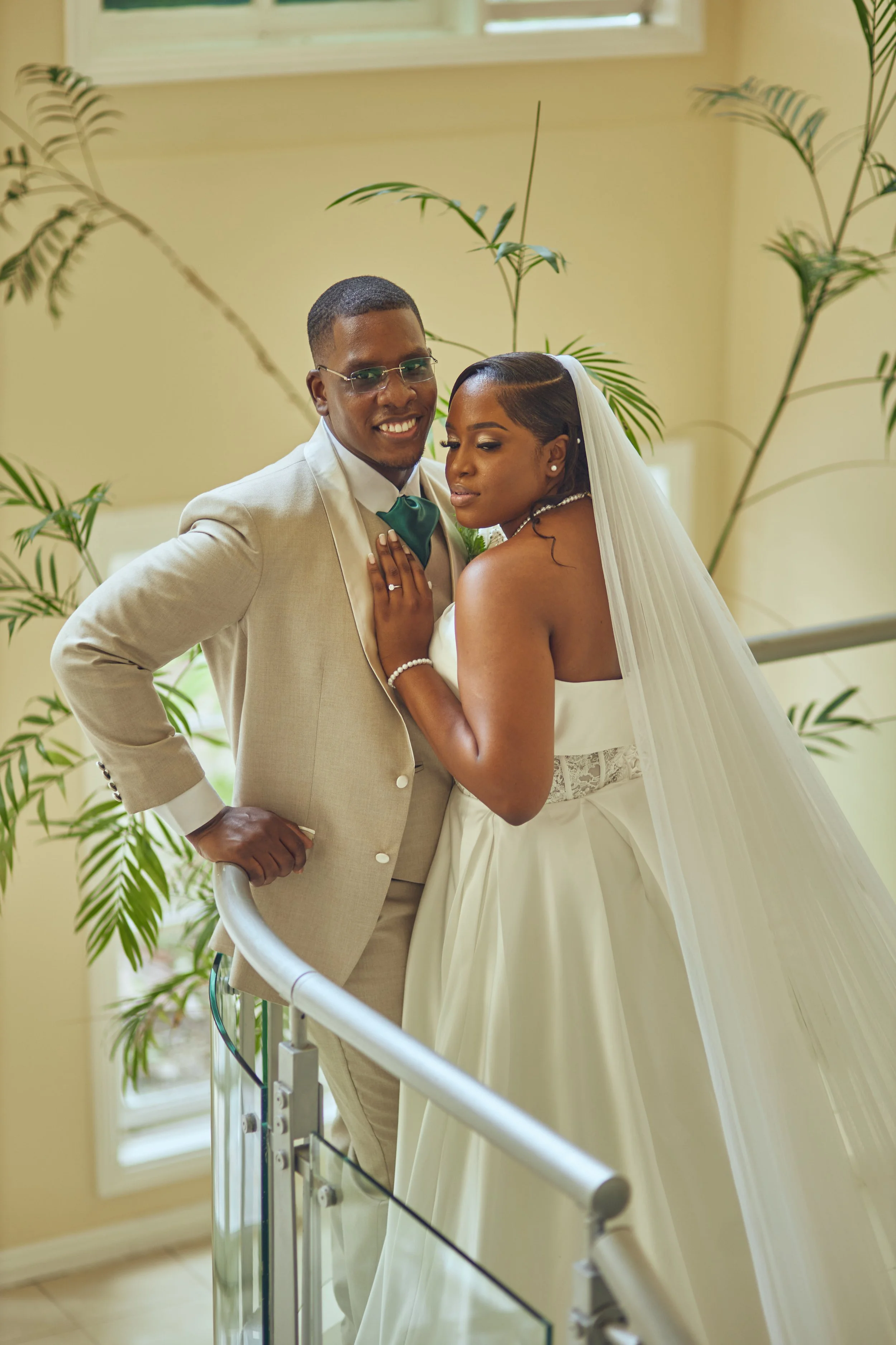 A newlywed couple in wedding attire, the groom in a beige suit with a teal bowtie and the bride in a strapless white wedding gown with a veil, posing indoors near green plants.