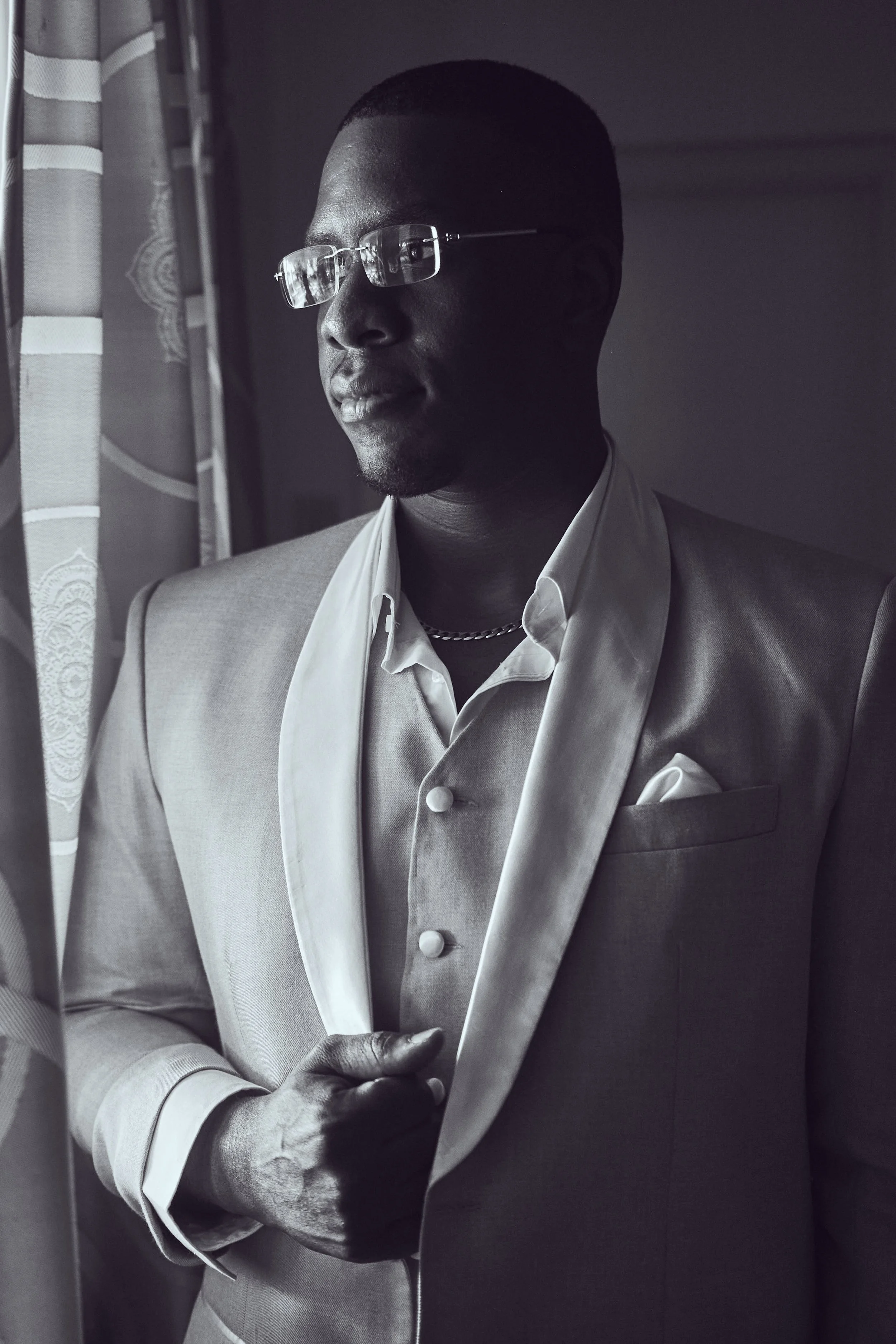 A man wearing glasses and a tuxedo looking out a window, holding the lapel of his jacket with one hand. The photograph is in black and white.