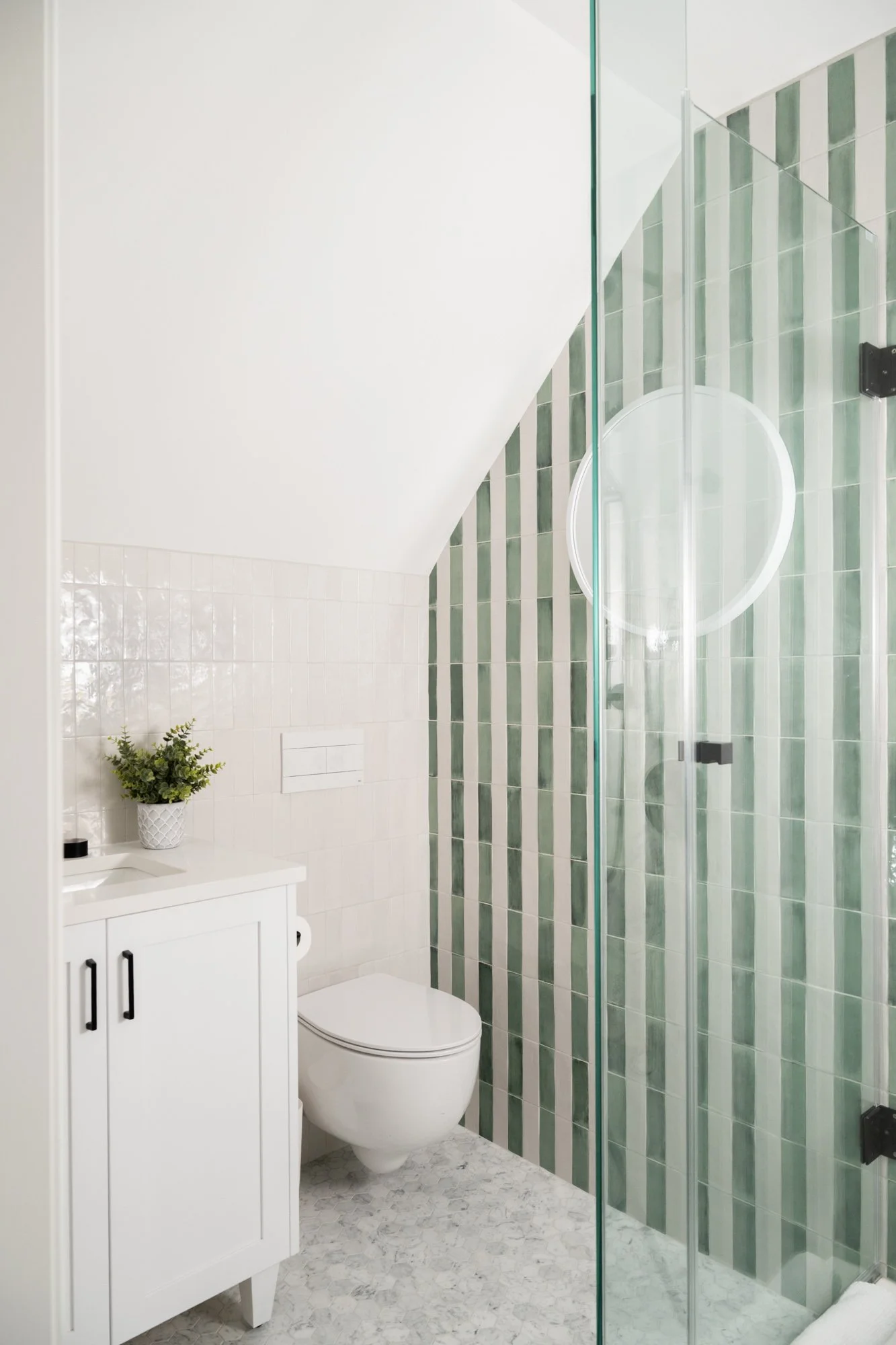 A small bathroom with a green and white striped wall tile, a circular mirror on the tile and a white vanity