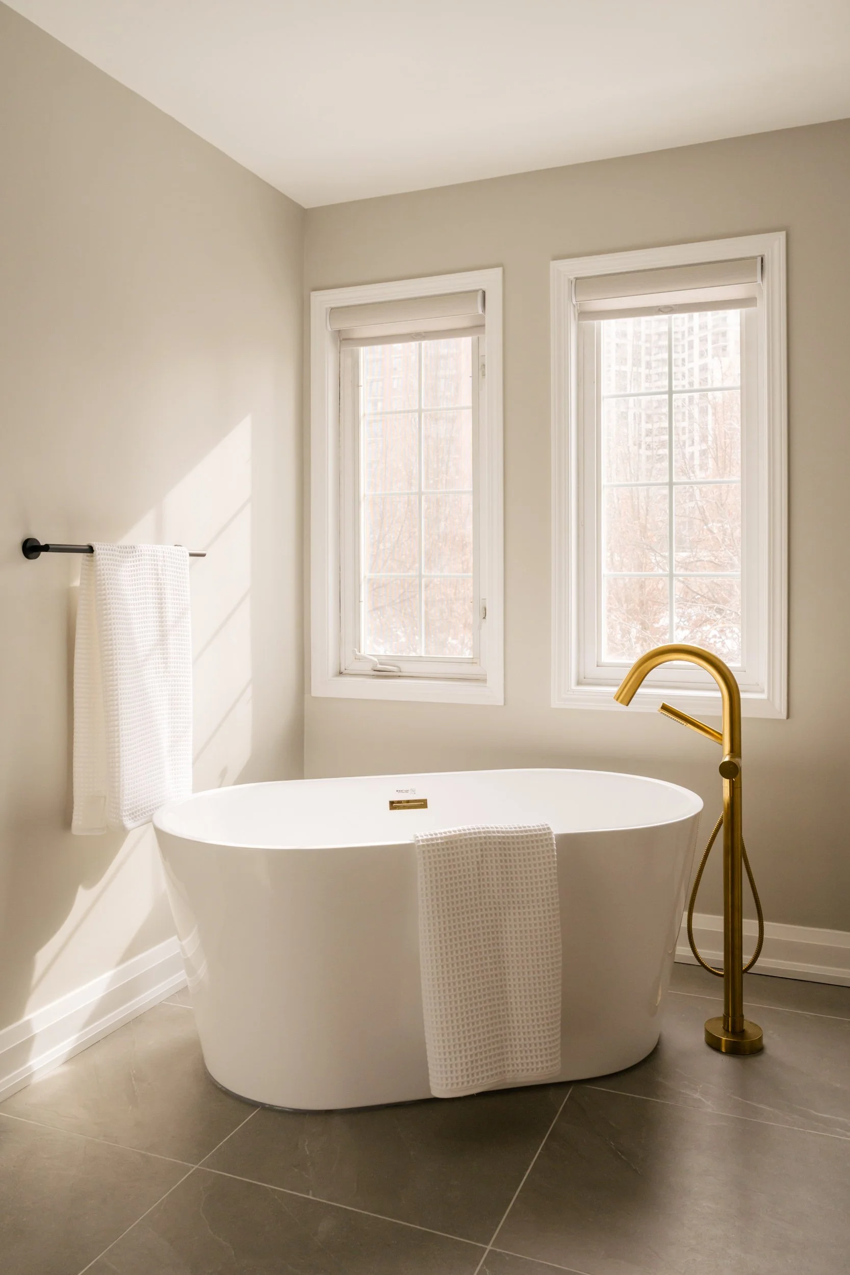 Freestanding tub with gold hardware in front of two windows.