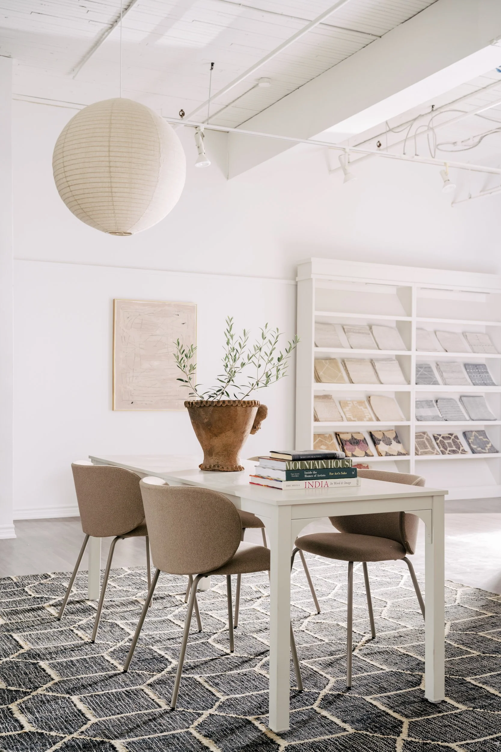 A meeting table with four chairs stand on top of a large patterned rug. A round paper lamp hangs above and a display case of rug samples stand in the background agains the wall.