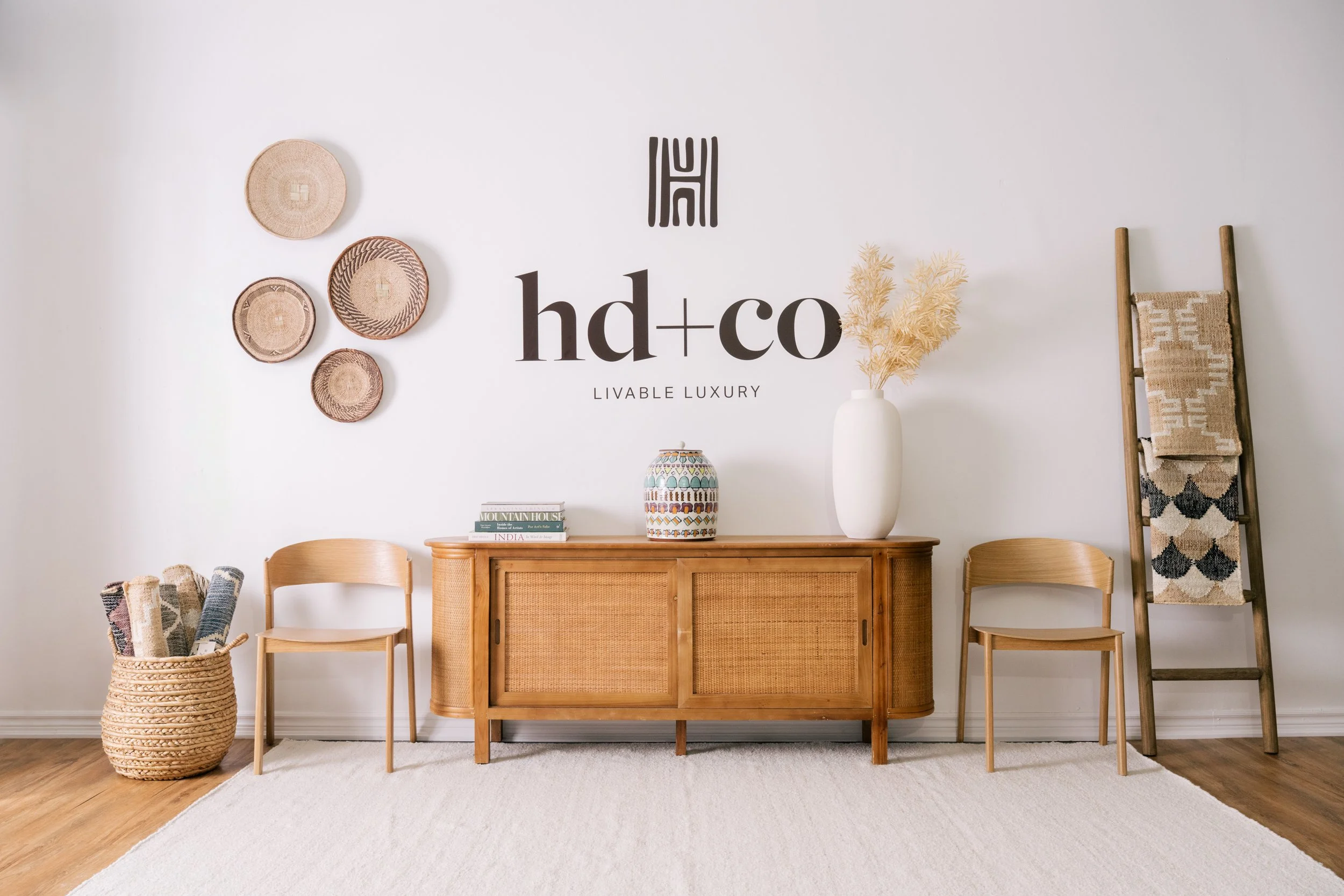 A credenza stands under the hd+co wall decal, surrounded by 2 chairs, all sitting on a large white rug. Decor is styled on top of the credenza.