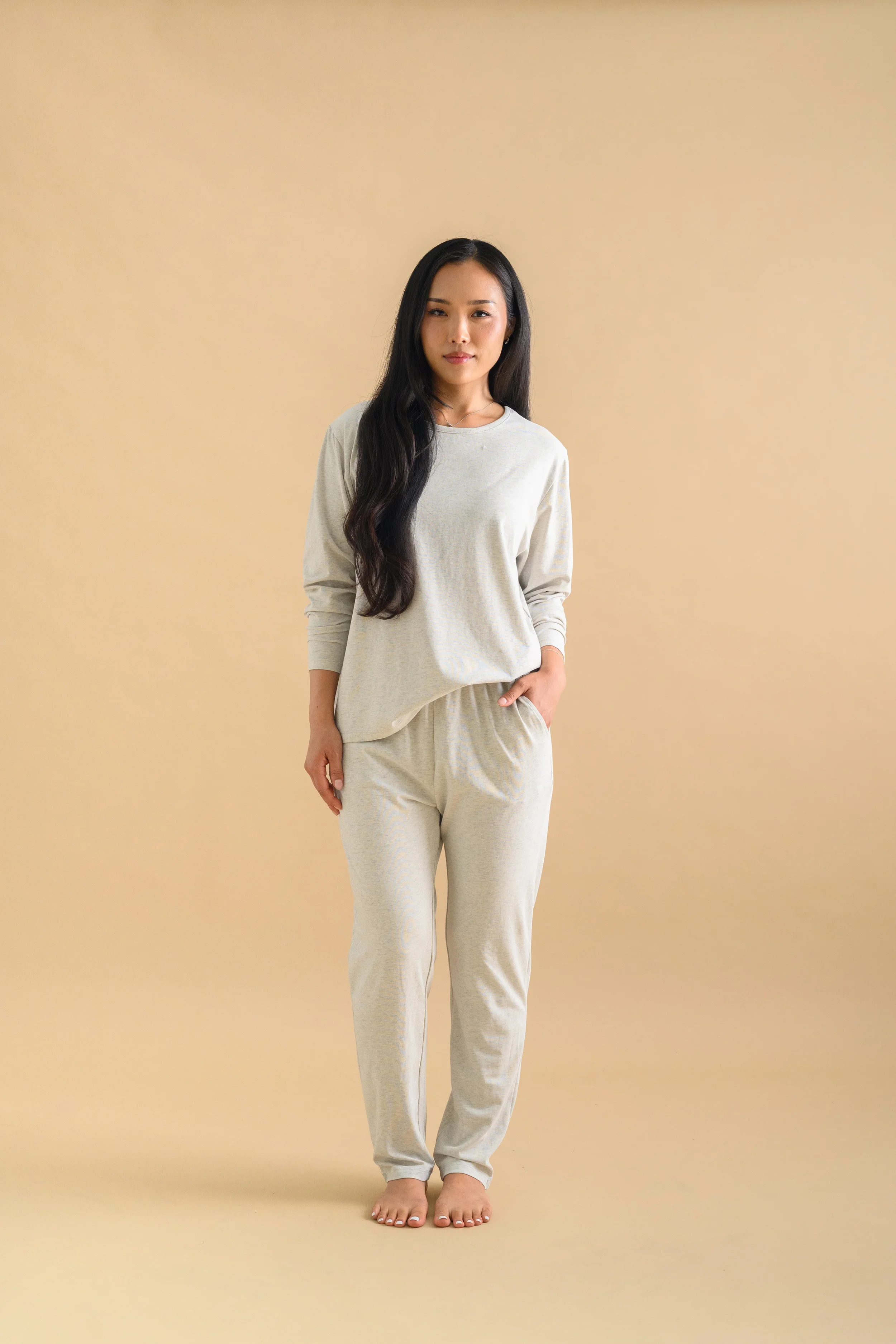 An Asian woman standing against a beige backdrop, wearing long sleeved shirt and long tapered pants by Heirloome House.