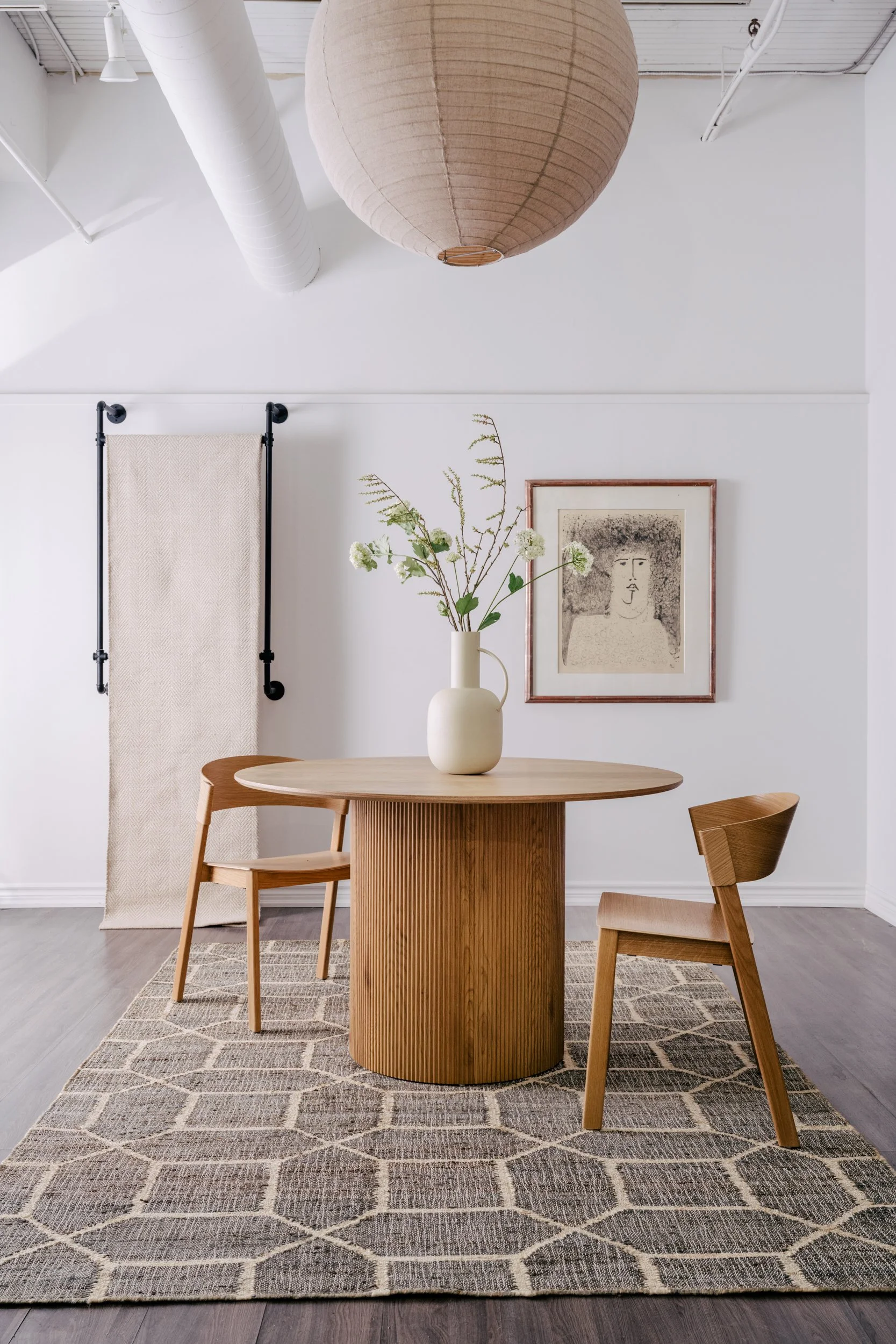A round wooden dining table with two chairs sit on top of a large patterned rug. A large paper lamp hangs above. An art frame hangs on the wall behind.