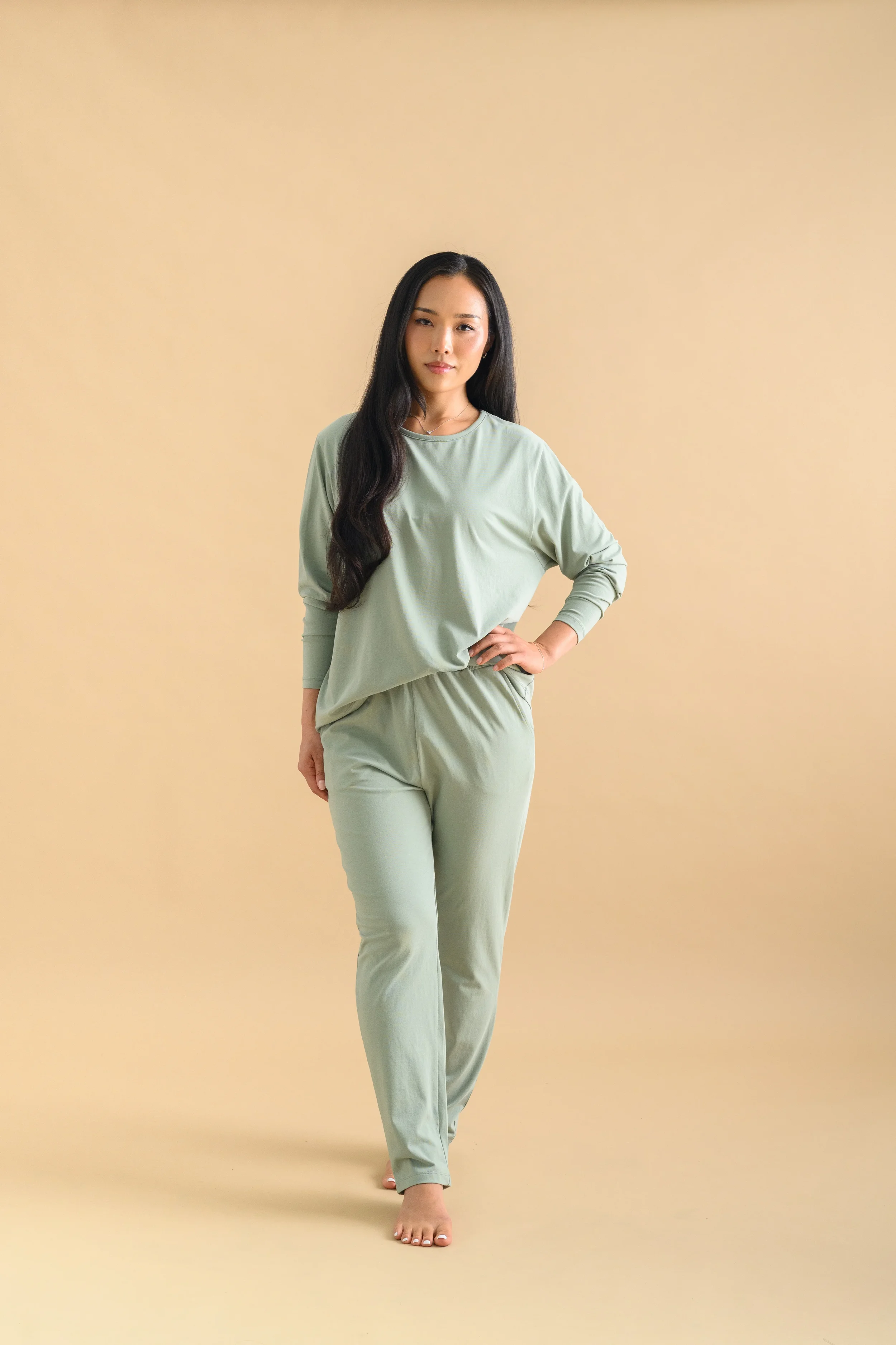 A woman standing against a beige backdrop, wearing a mint green long sleeved top and tapered pants by Heirloome House.
