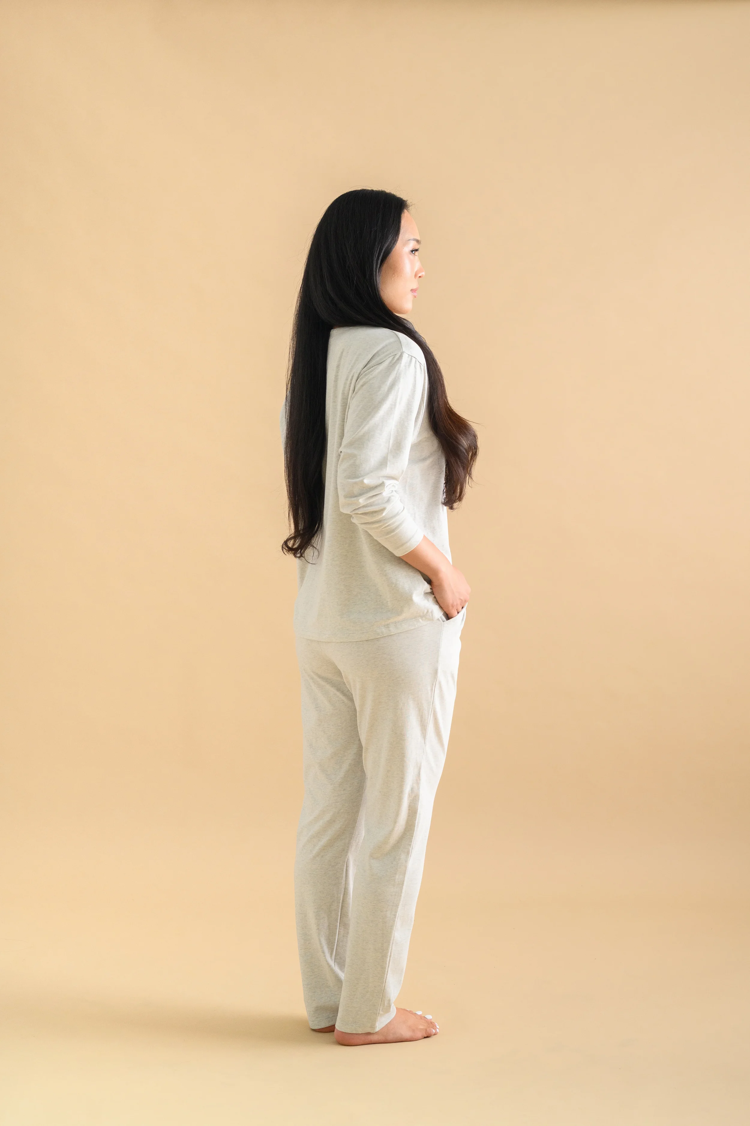 Side profile of an Asian woman standing against a beige backdrop, wearing long sleeved shirt and long tapered pants by Heirloome House.