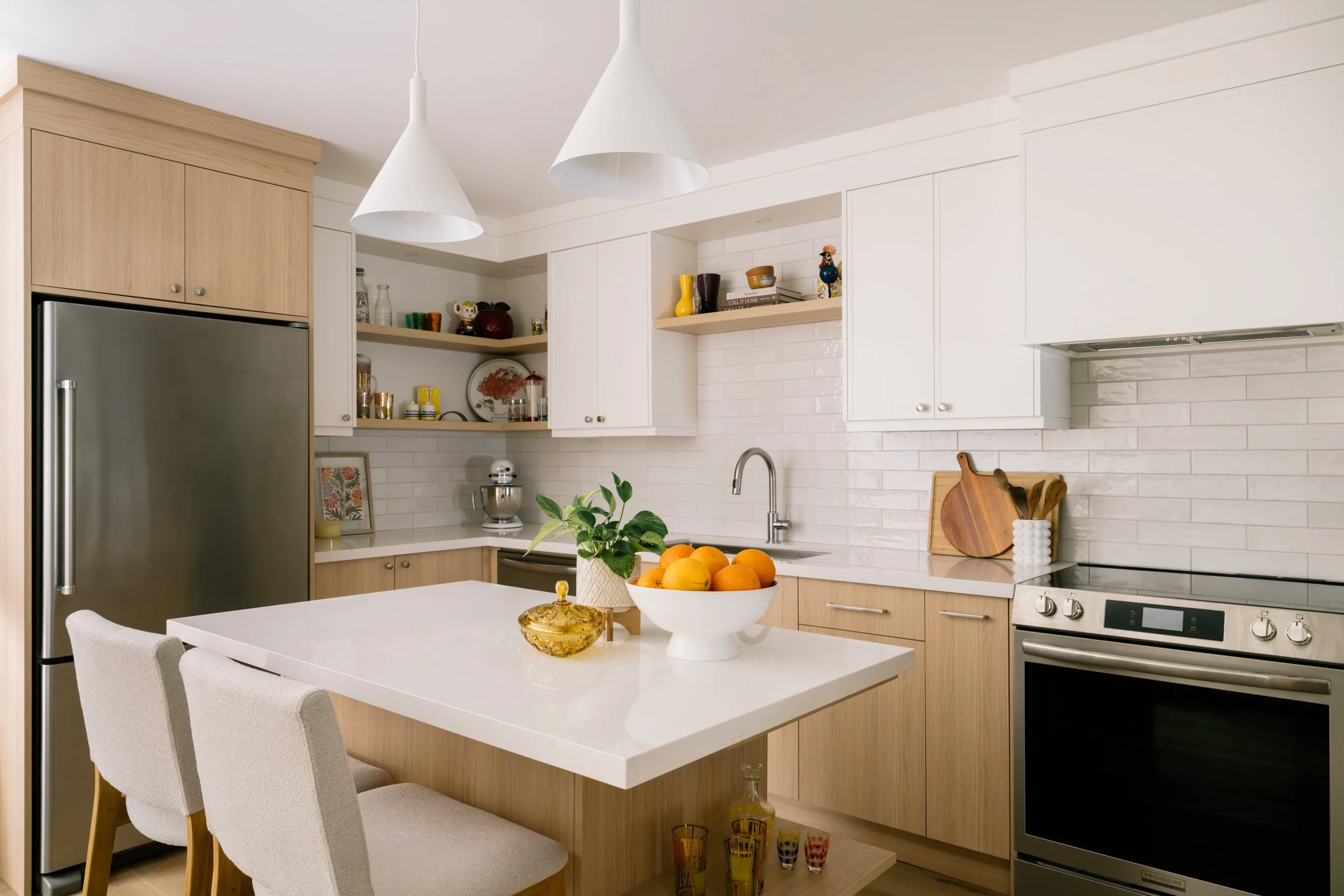 A kitchen with white upper cabinets and light wood lower cabinets. A small kitchen island in the middle of the kitchen with two stools and two pendants lights above.