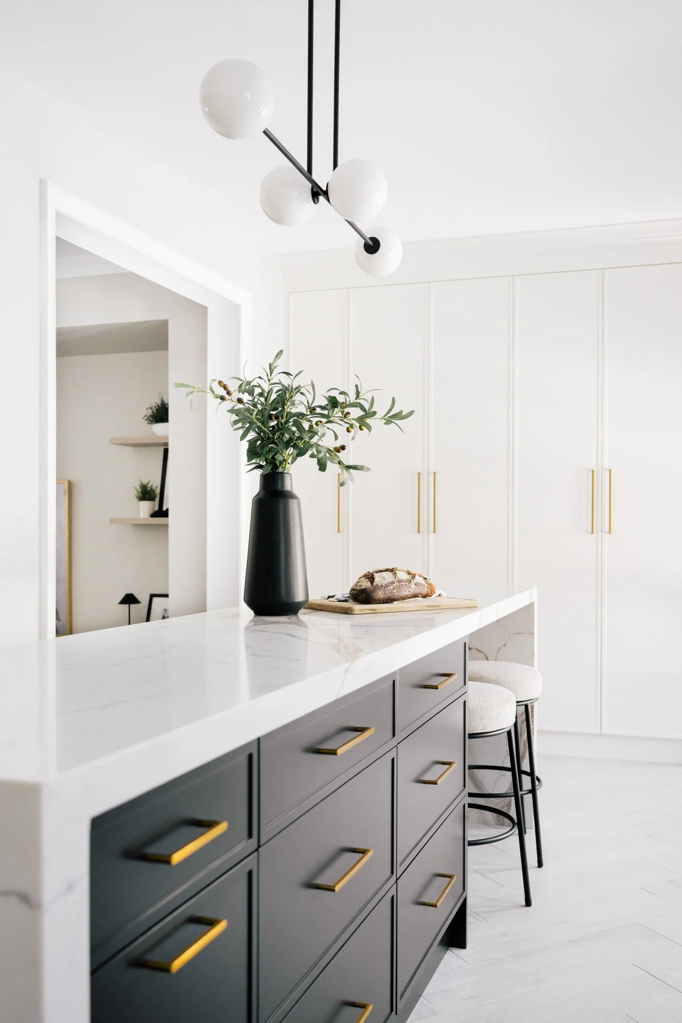 A white marbled waterfall kitchen island with black lower cabinets and gold handles. A black vase and loaves of bread sit on the island.