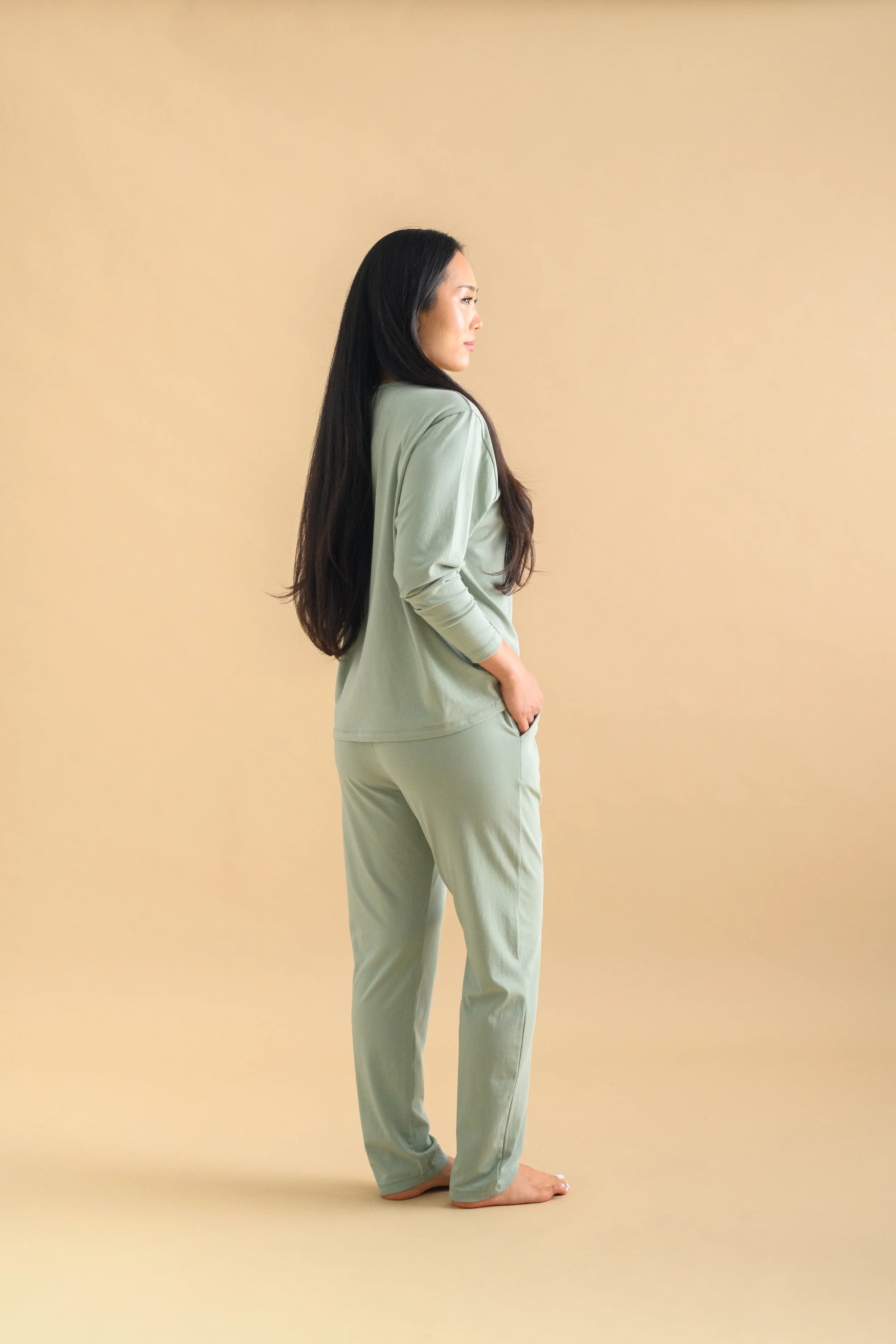A side view of a woman standing against a beige backdrop, wearing a mint green long sleeved top and tapered pants by Heirloome House.
