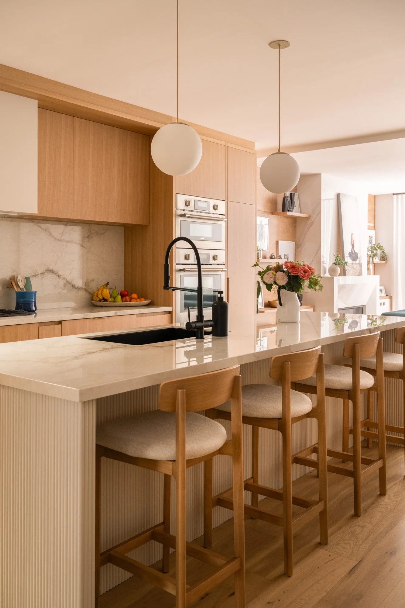 A large open kitchen with light wood cabinets and marble countertop and backsplash with a long kitchen island and four tall stools.
