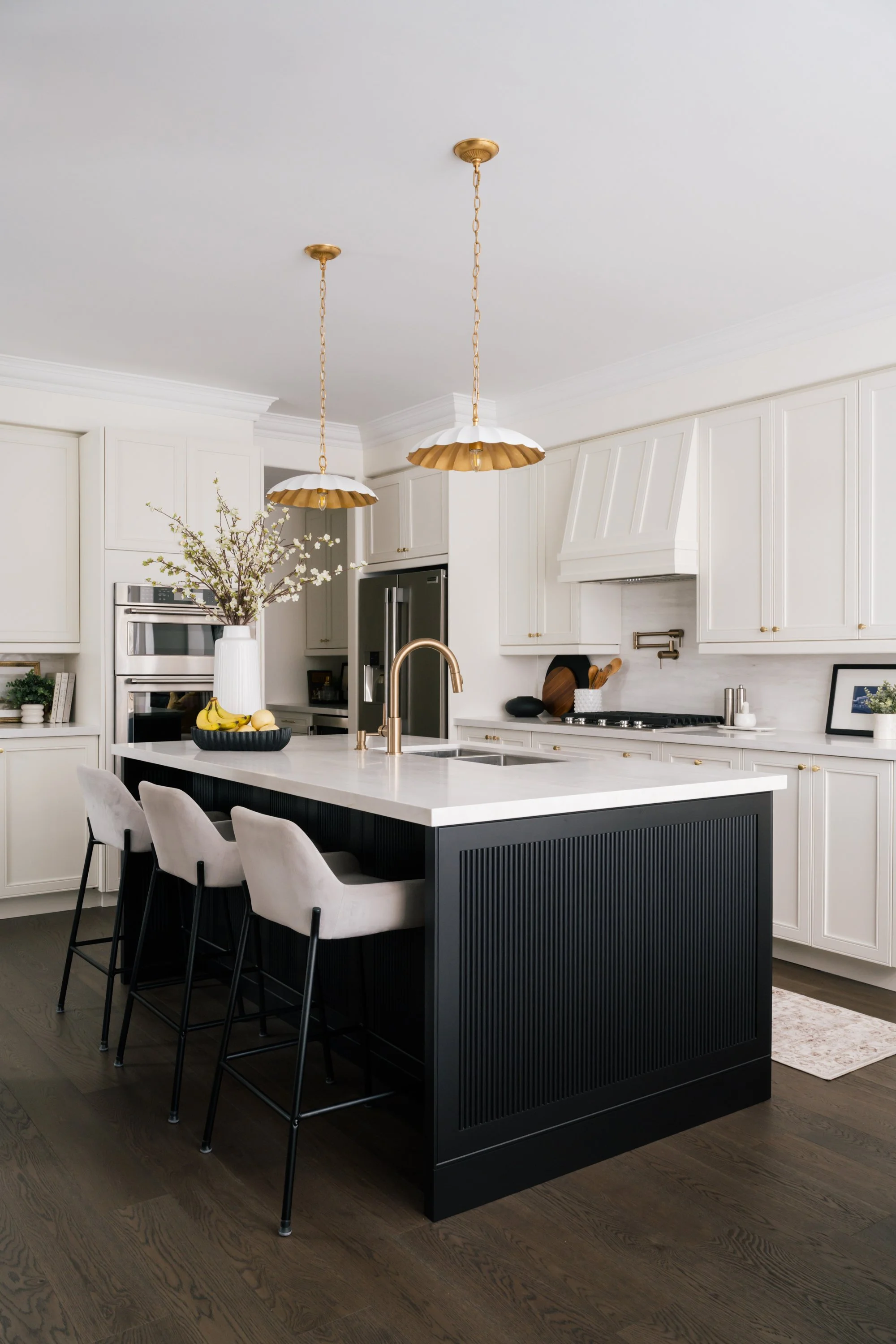Off-white L-shaped kitchen and a large island with black lower cabinets. Two scalloped pendants hang above.