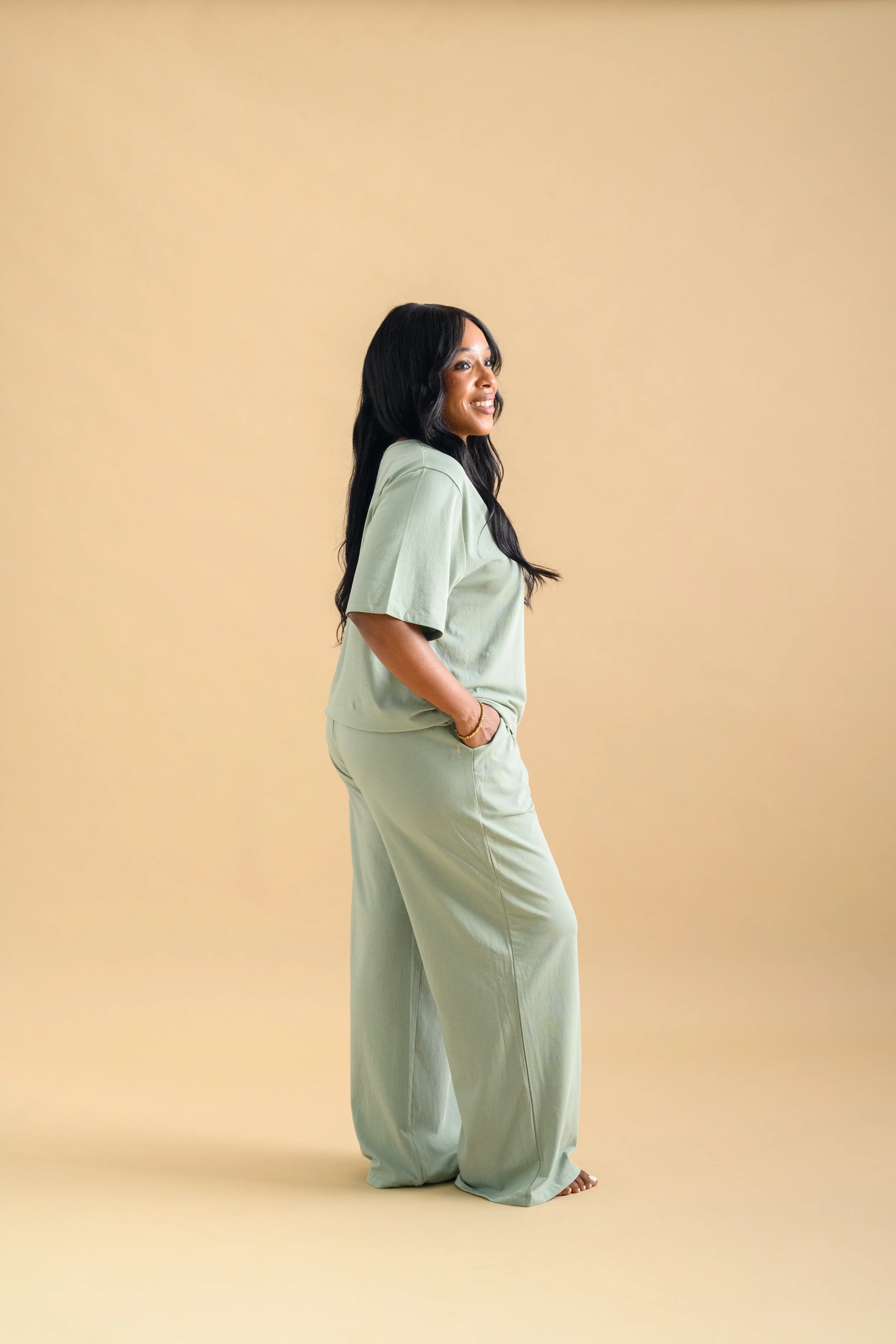 A woman standing against a beige backdrop, wearing a mint green short sleeved shirt and long loose pants by Heirloome House.