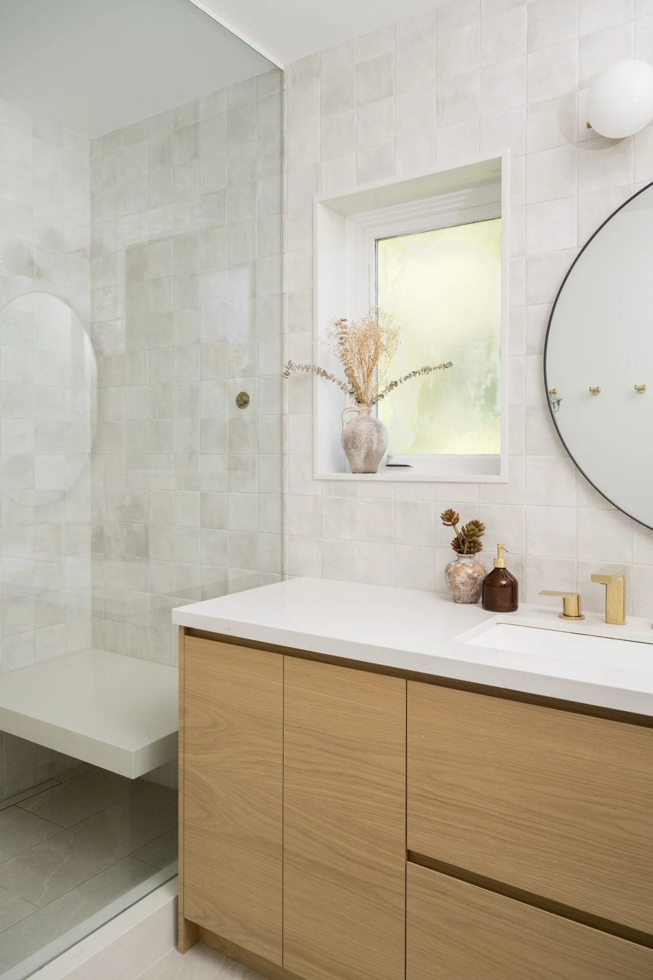 Primary bathroom vanity with square tile backsplash, gold hardware, and circular mirror. A shower with a glass door and bench beside the vanity.