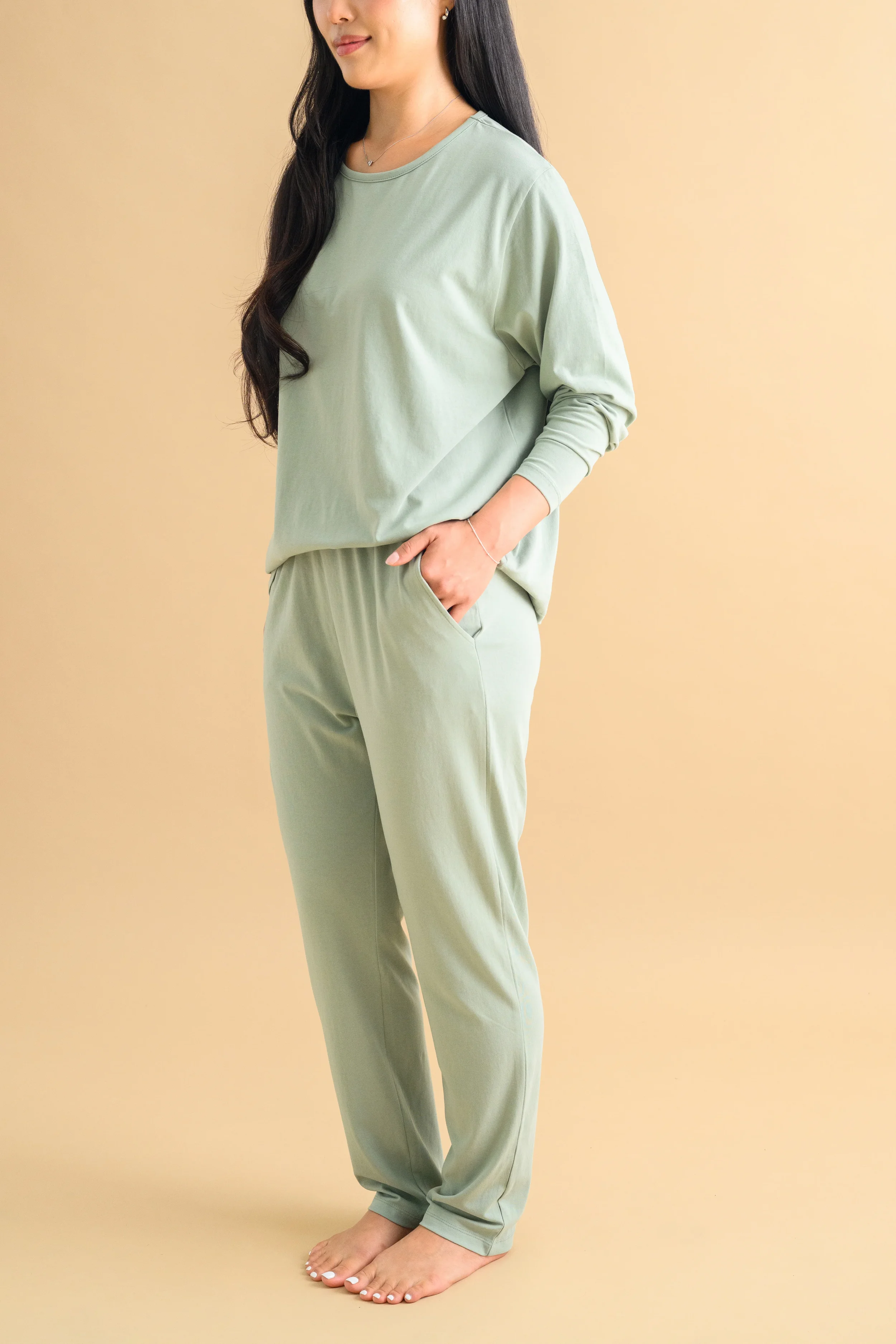 A woman standing against a beige backdrop, wearing a mint green long sleeved top and tapered pants by Heirloome House.