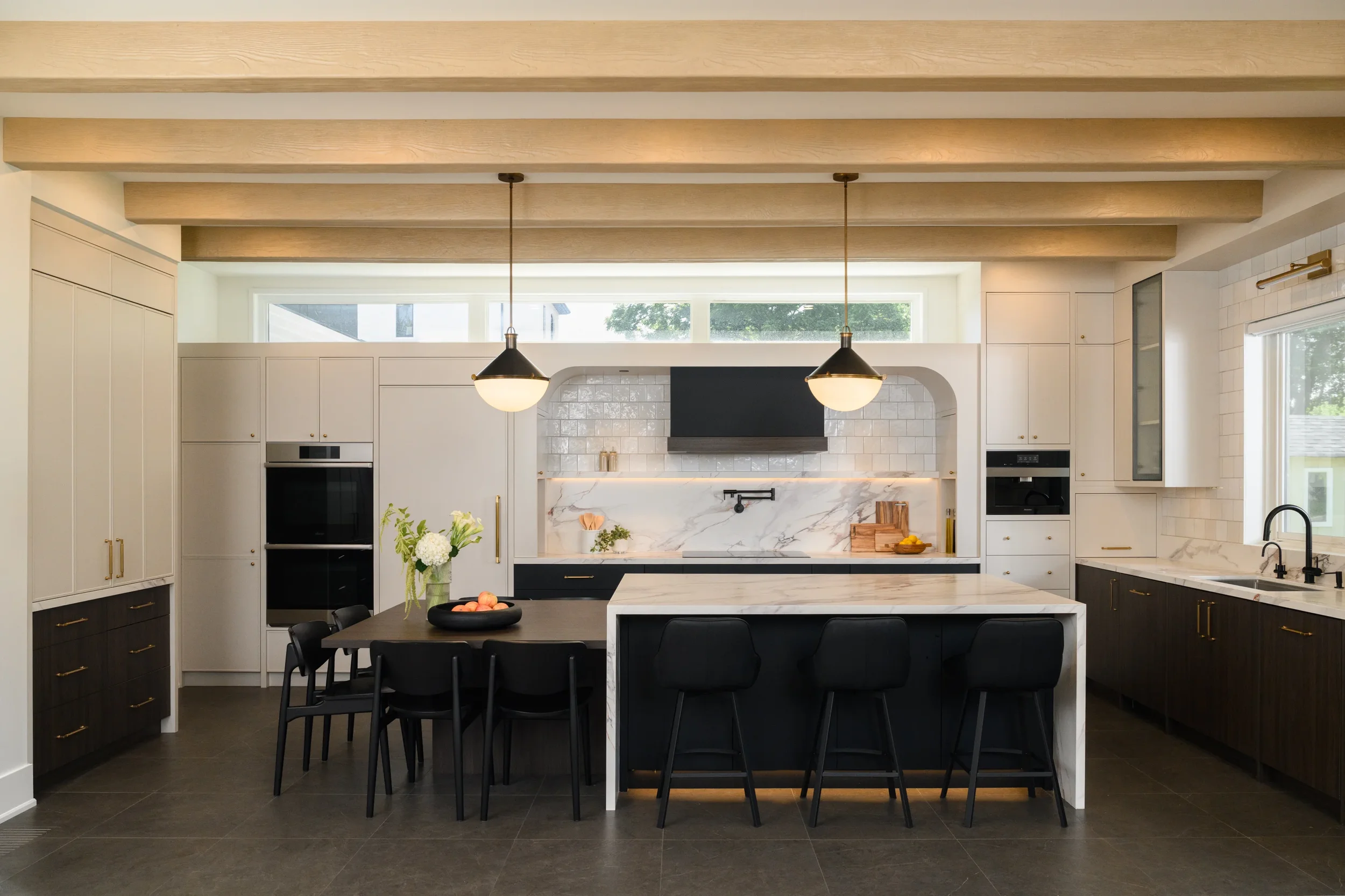 A large black and white kitchen with wood beams in the ceiling and additional windows above the stove area. The kitchen lights are turned on to create warm lighting throughout the space.