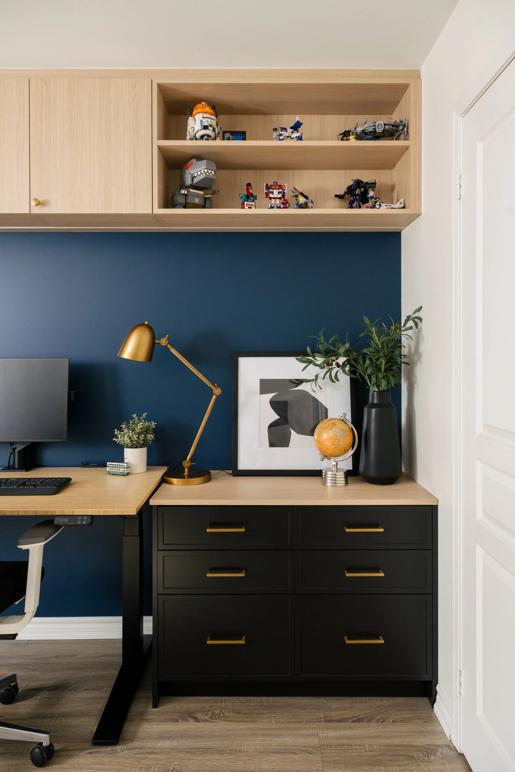 Home office with built-in shelves. Open shelving above displays toy figurines. The wall behind the desk is painted blue.