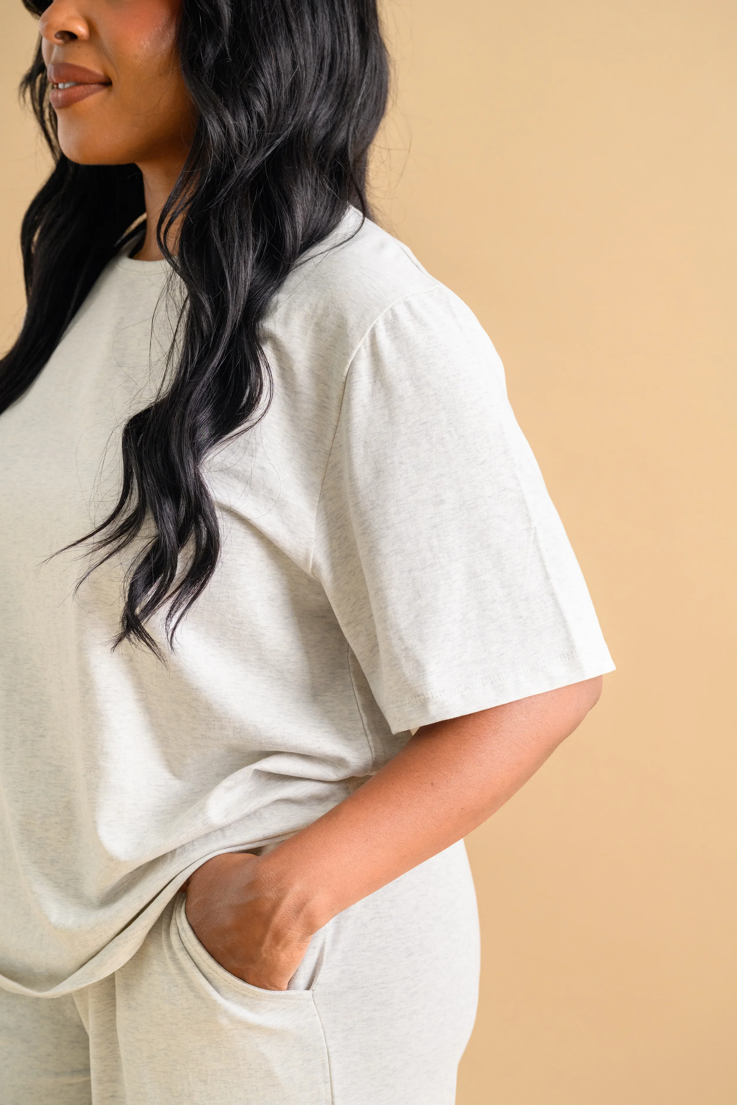 A close up of a woman standing against a beige backdrop, wearing an oat-coloured long sleeved top and tapered pants by Heirloome House.