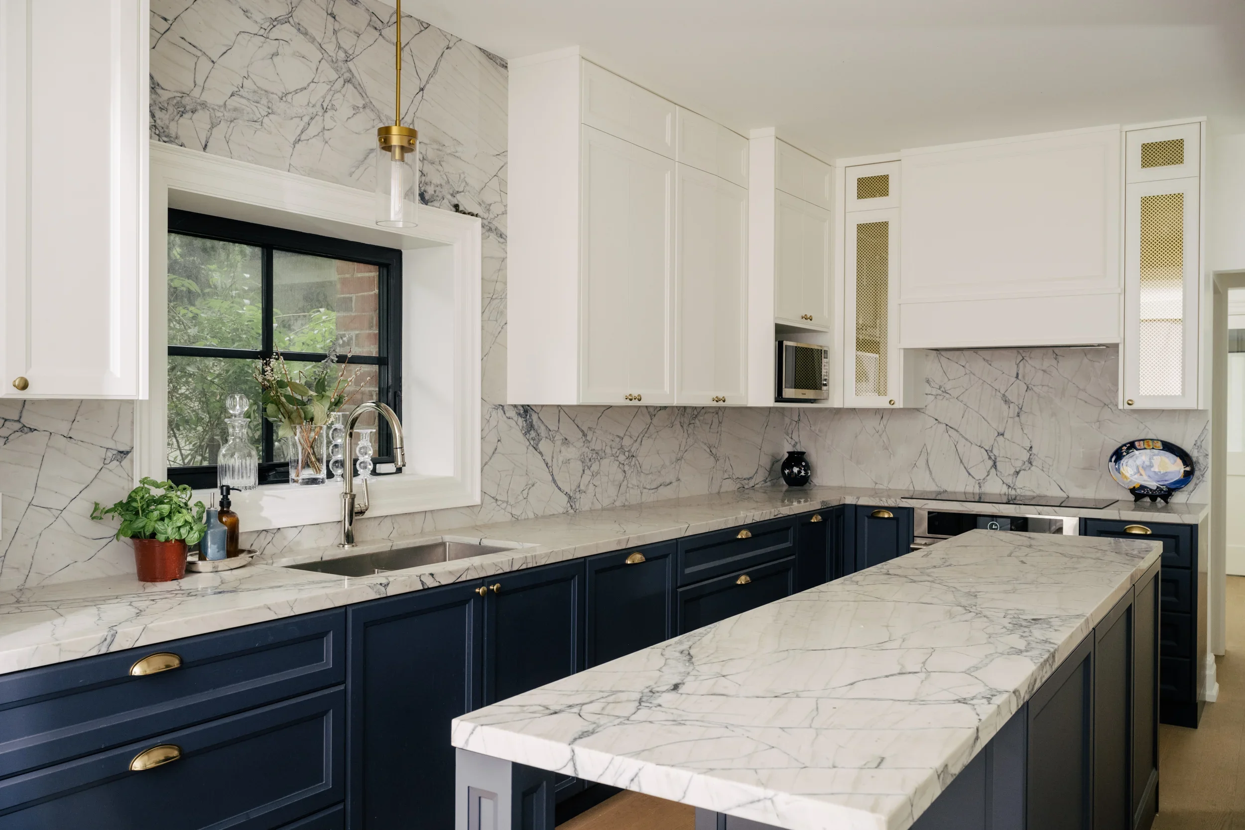 A long kitchen with marble backsplash and navy blue lower kitchen cabinets and gold handle pulls