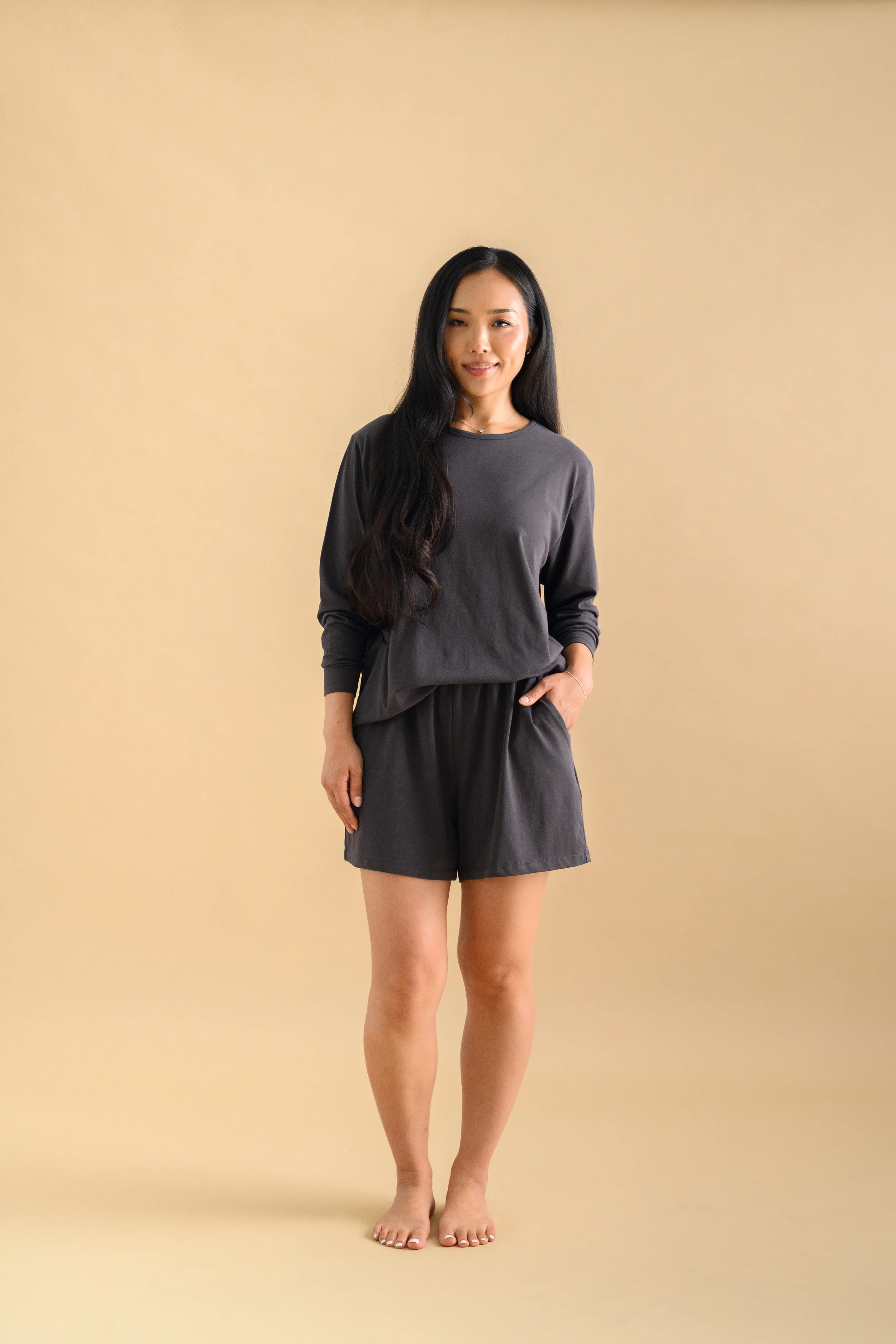 An Asian woman standing against a beige backdrop, wearing long sleeved shirt and shorts by Heirloome House.