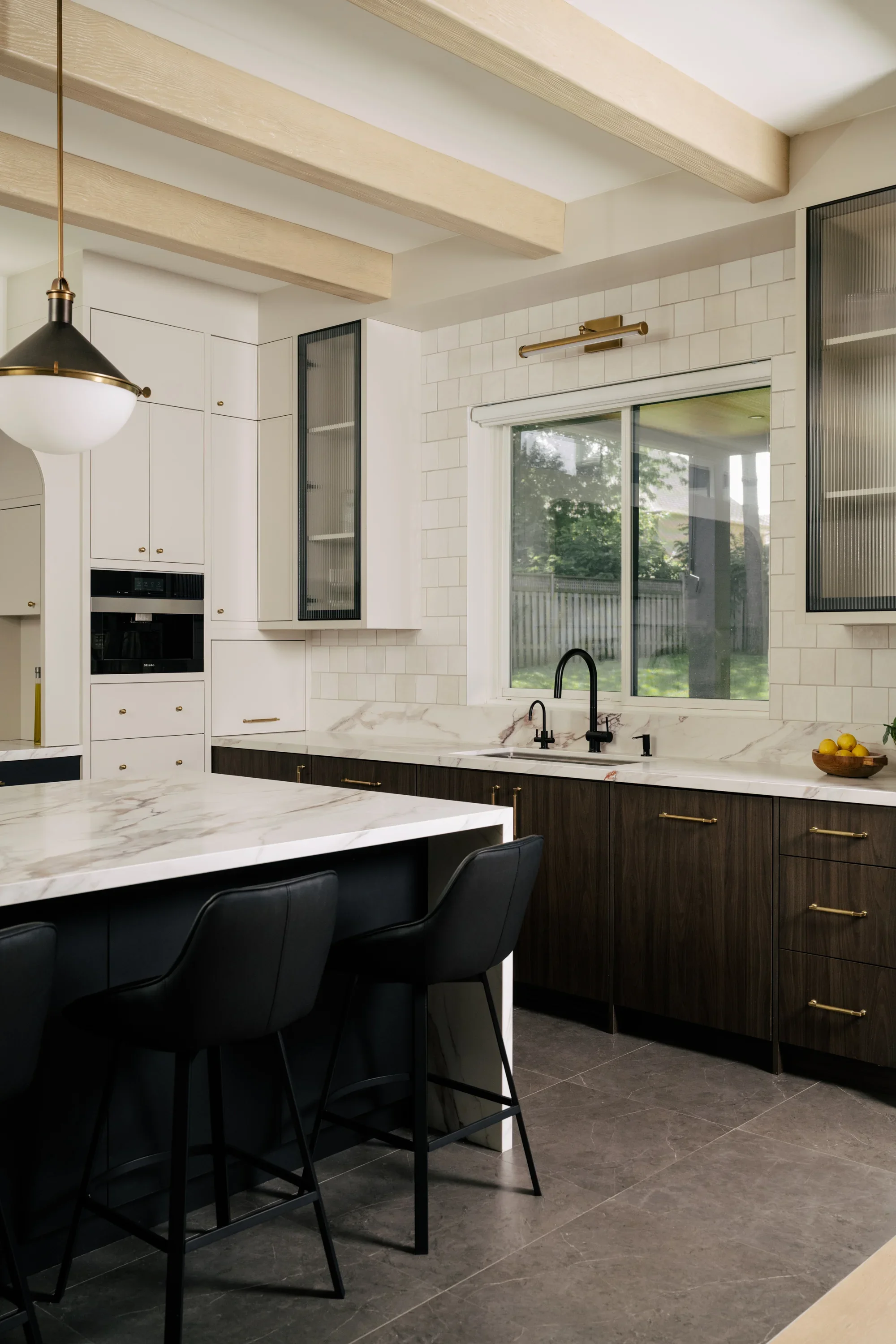 A view of a kitchen sink with a large window looking out into the backyard. In the foreground is a large kitchen island with black stools.