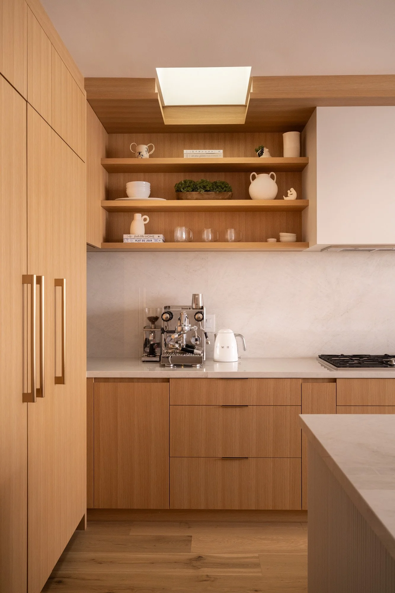 A kitchen with light, warm wood cabinets, an open shelf for cups and an espresso machine on the countertop. A small skylight allows sunlight from above.