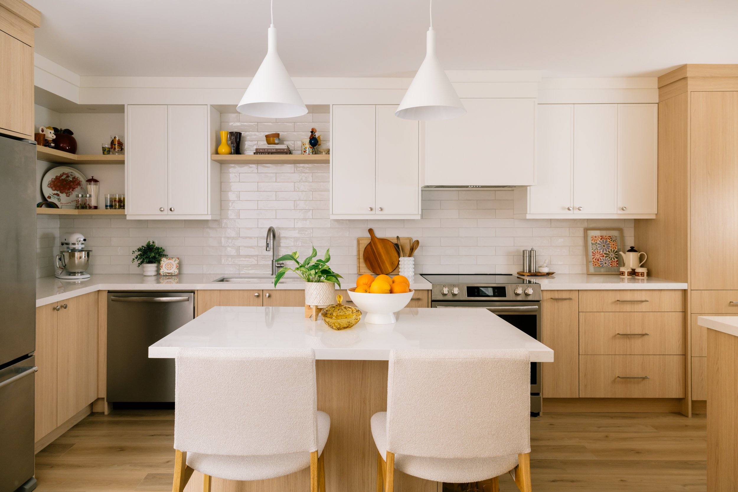 A kitchen with white upper cabinets and light wood lower cabinets. A small kitchen island in the middle of the kitchen with two stools and two pendants lights above.