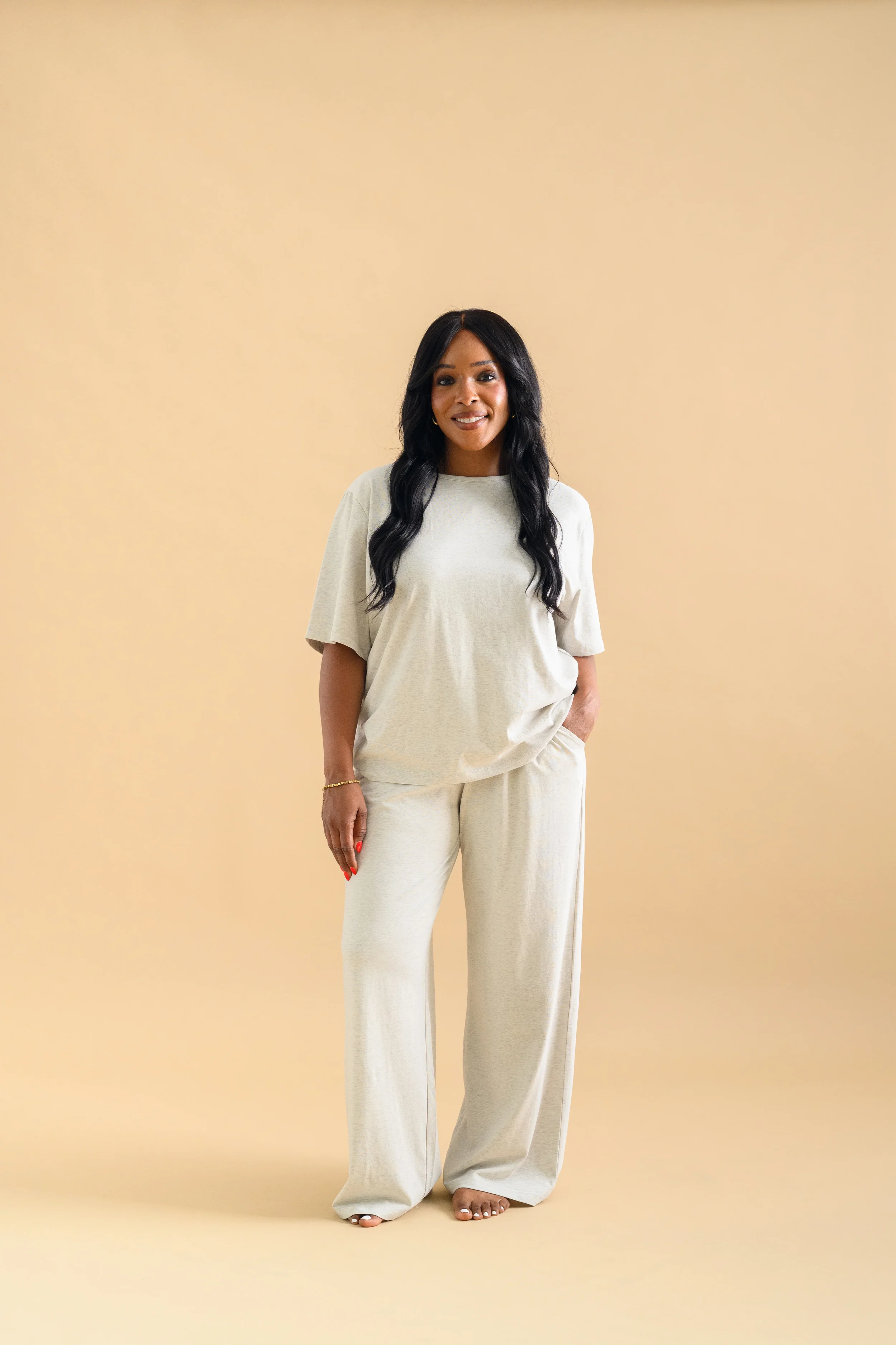 A woman standing against a beige backdrop, wearing an oat-coloured long sleeved top and tapered pants by Heirloome House.