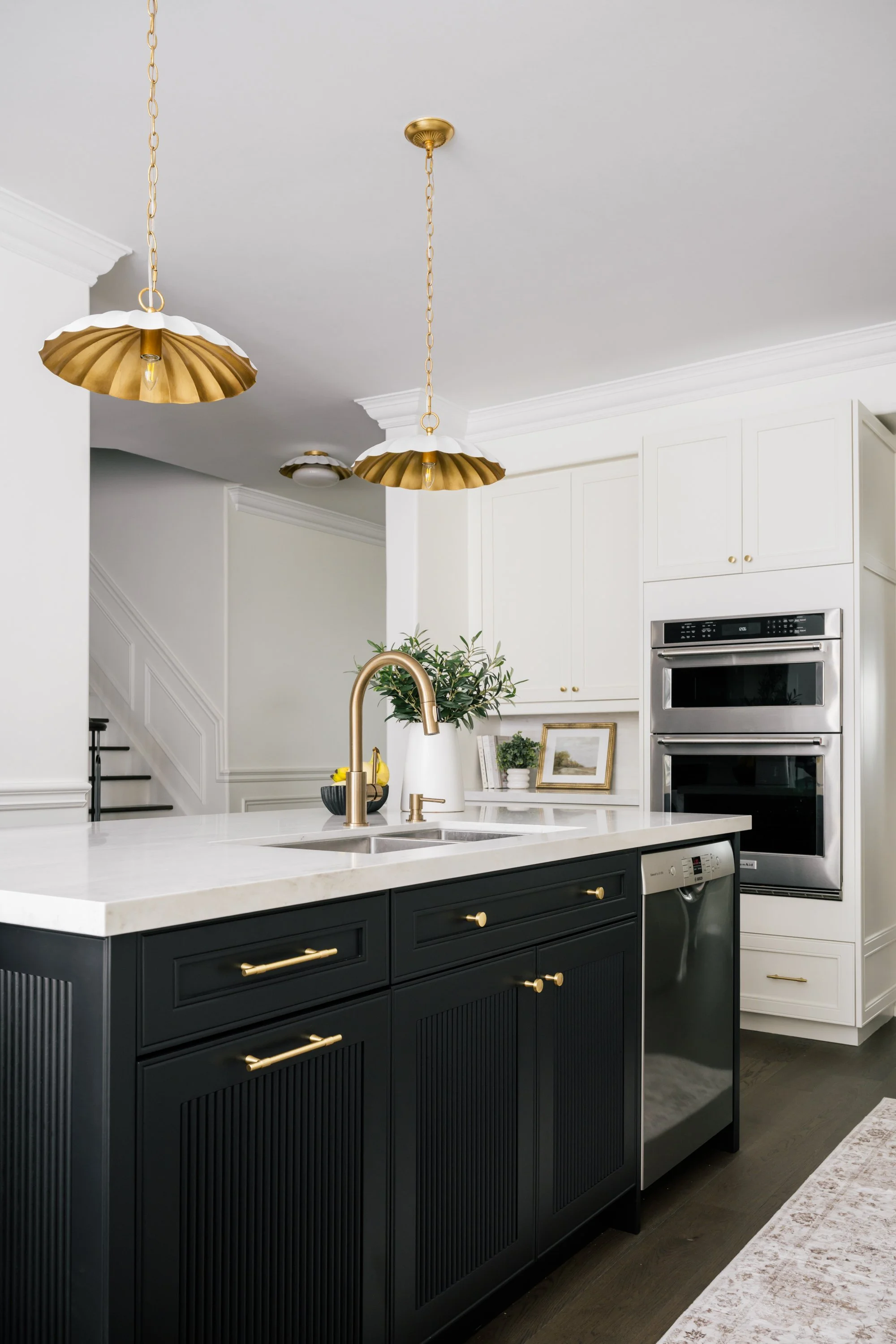 Off-white L-shaped kitchen and a large island with black lower cabinets. Two scalloped pendants hang above. An integrated oven and stove are built into one side of the kitchen.