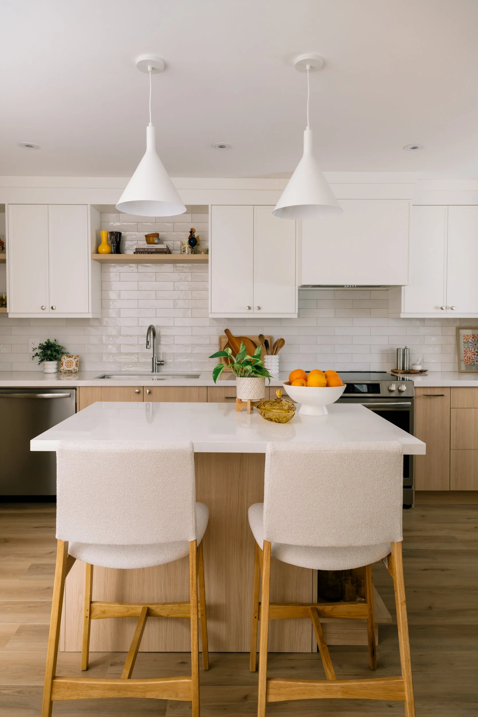 A kitchen with white upper cabinets and light wood lower cabinets. A small kitchen island in the middle of the kitchen with two stools and two pendants lights above.