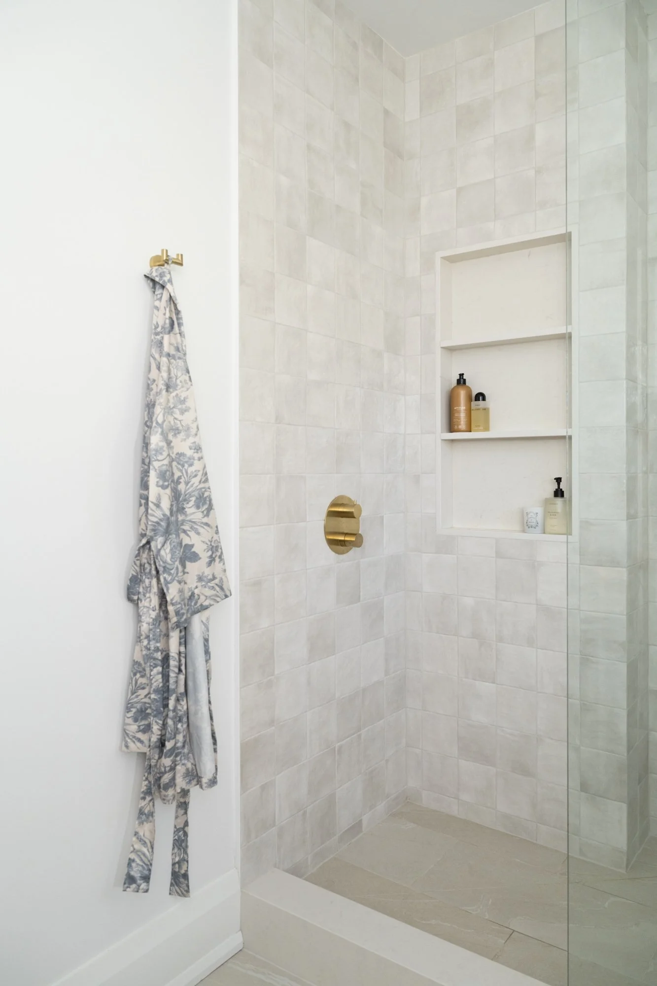 A shower with square tiles and a 3-level wall niche. A floral robe hangs on the wall.