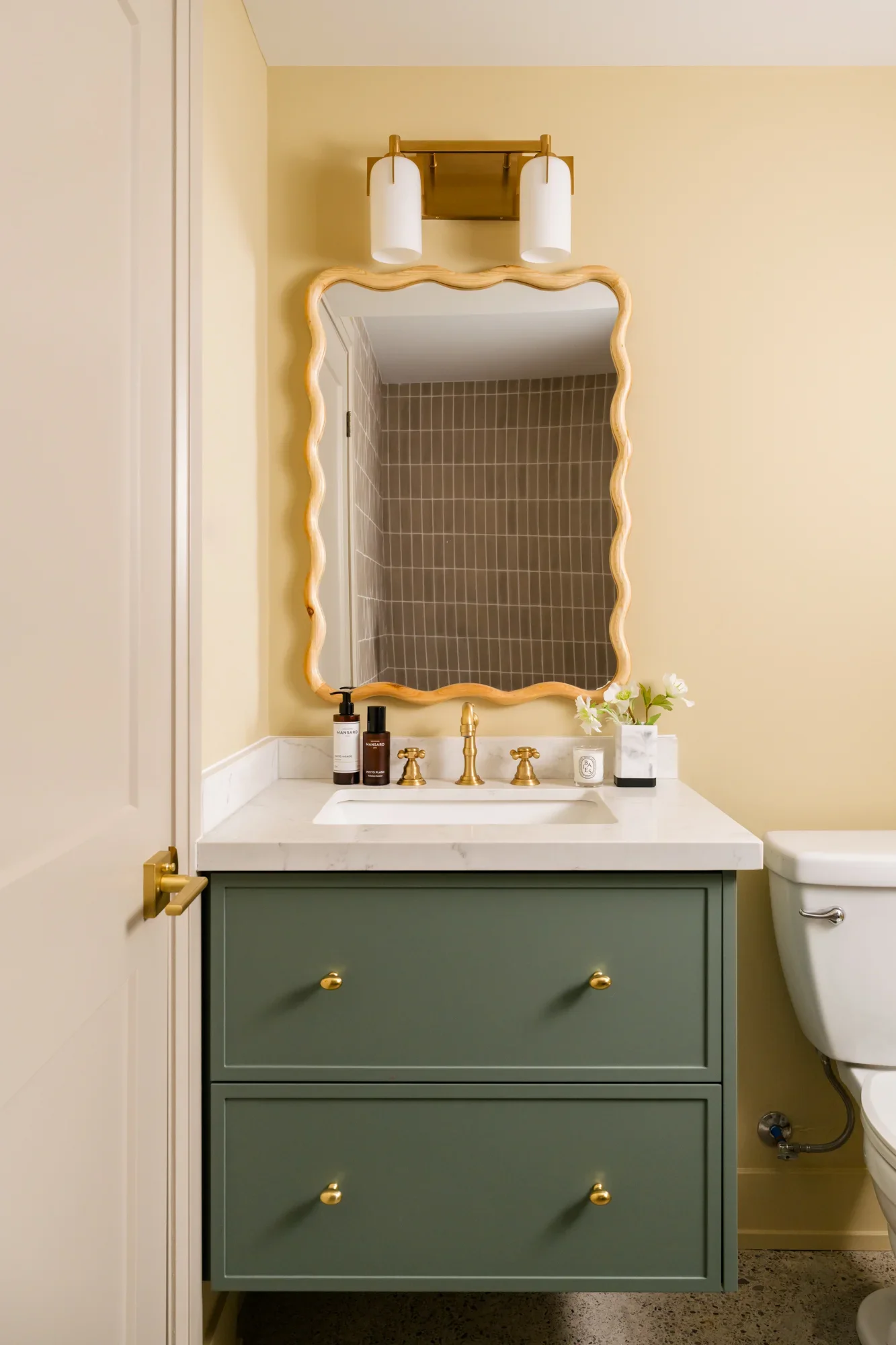 Basement bathroom vanity. The mirror has a whimsical squiggly frame. The cabinet is a soft sage green and brushed gold hardware.