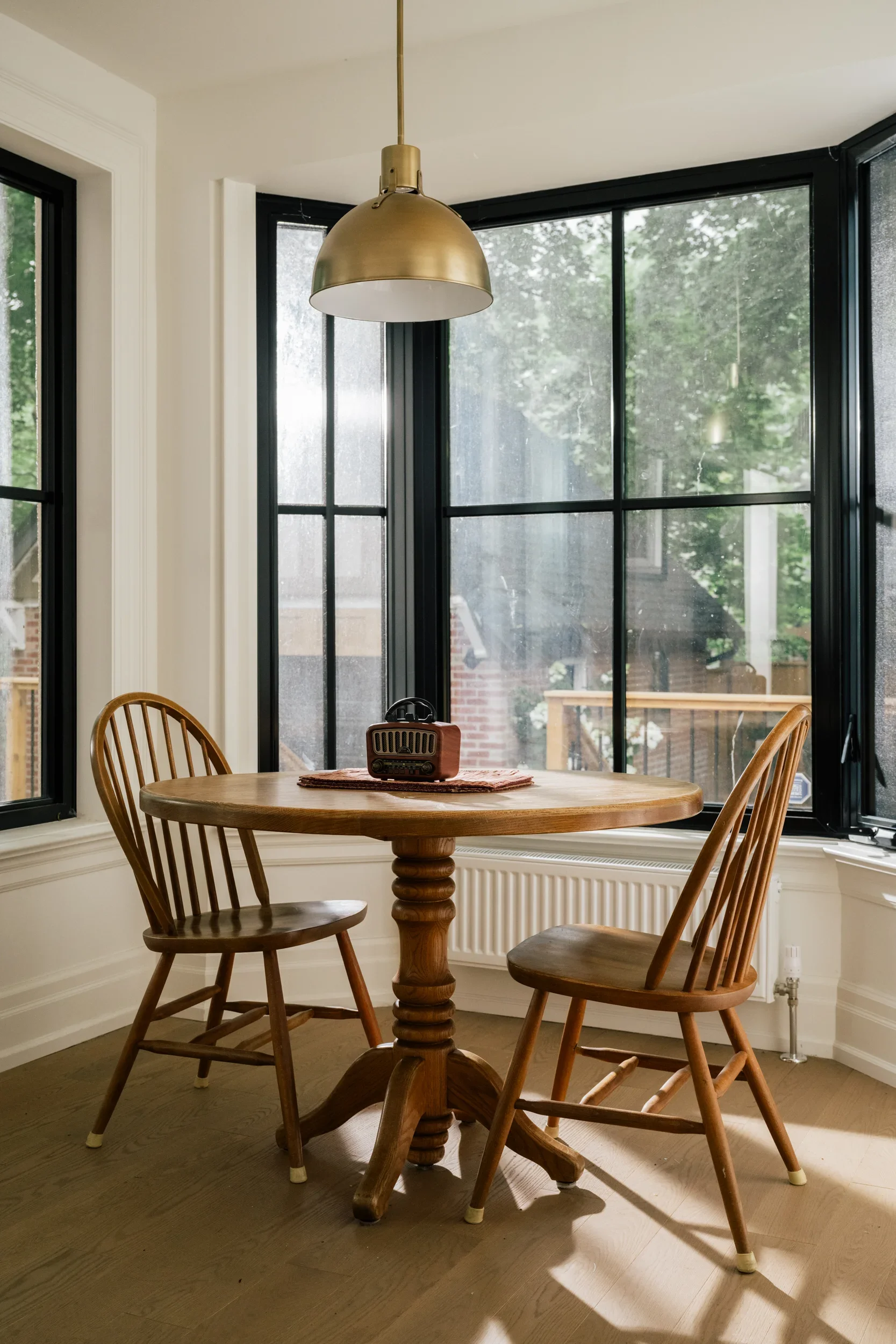 A wooden dining table and two chairs in front of a black bay window with light streaming in through the window