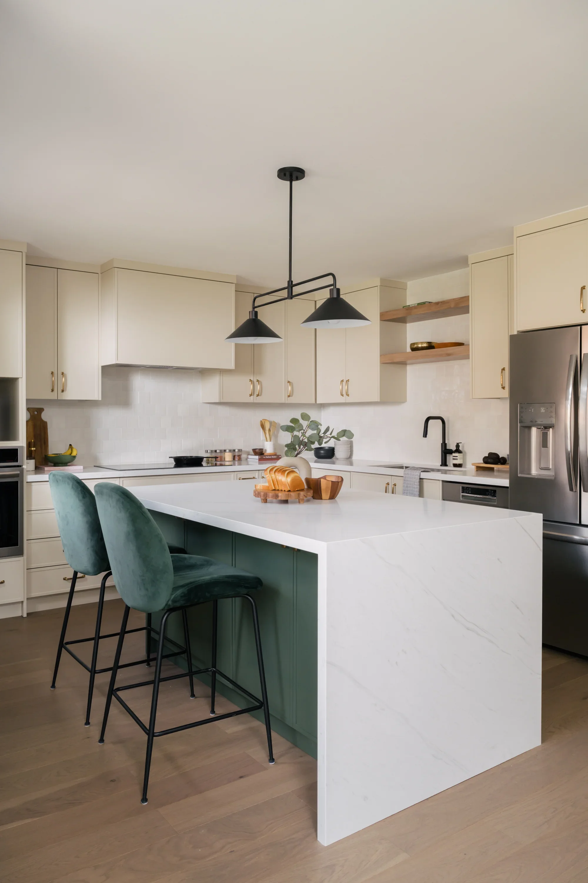 A full top to bottom view of a cream-coloured kitchen with a large kitchen island and green velvet stools. Slices of bread on a wood board and vase are styled on the island.