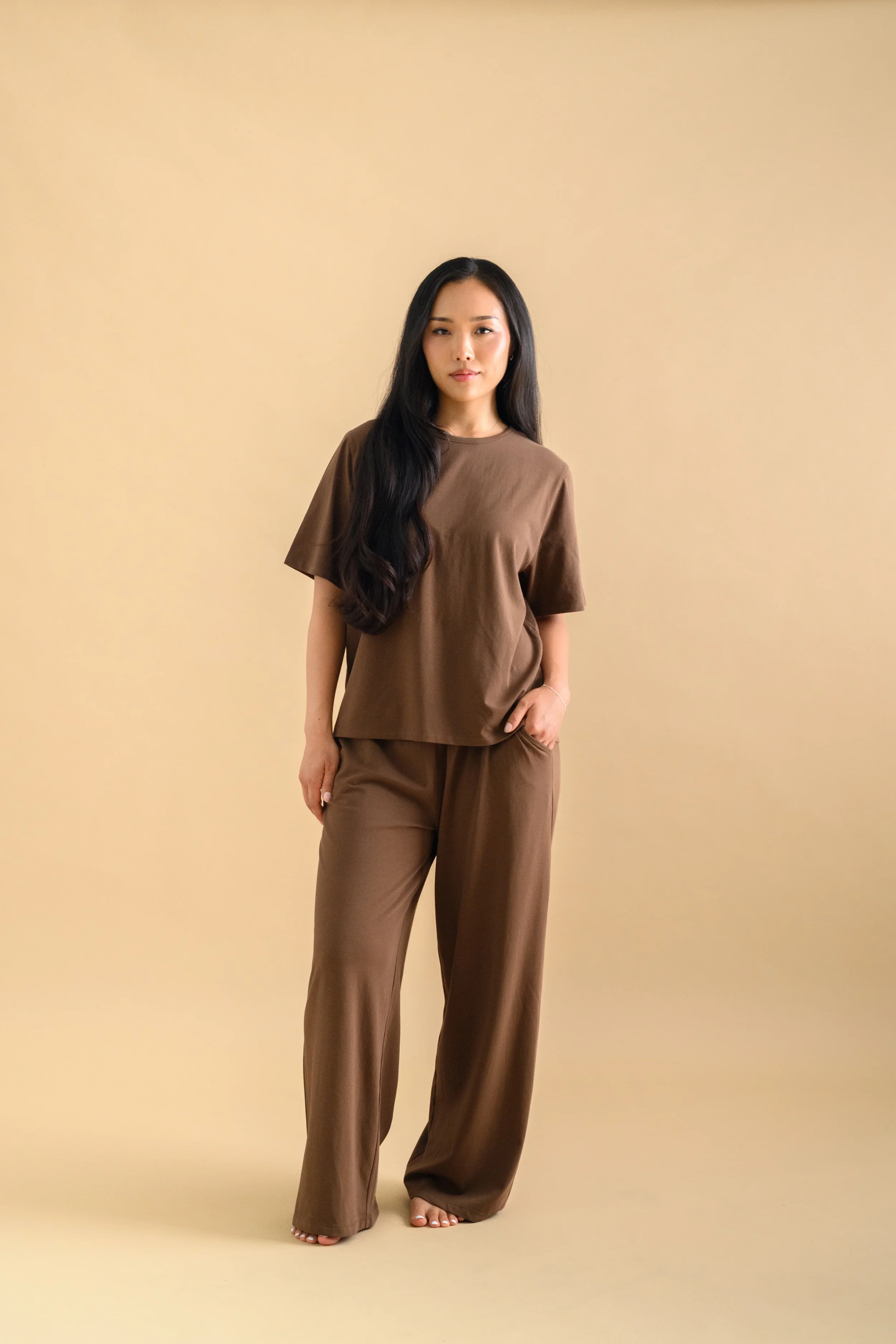 A woman standing against a beige backdrop, wearing a brown short sleeved shirt and long loose pants by Heirloome House.