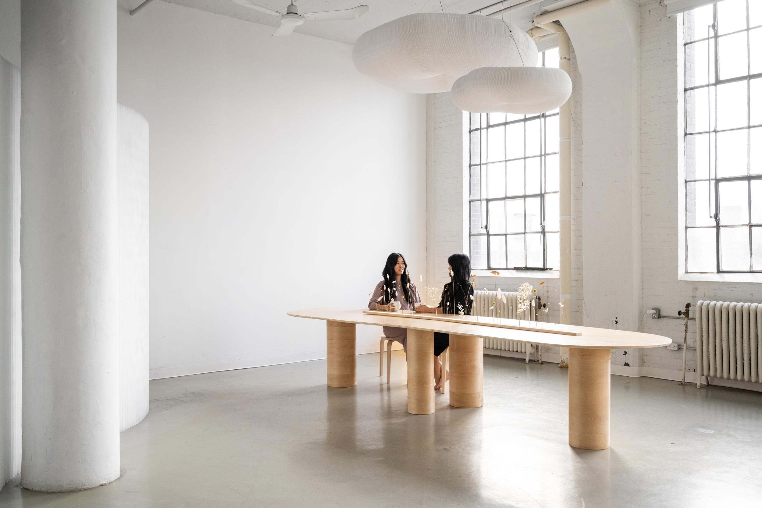 Two young women having coffee at a long wooden table in a large studio space with large windows.