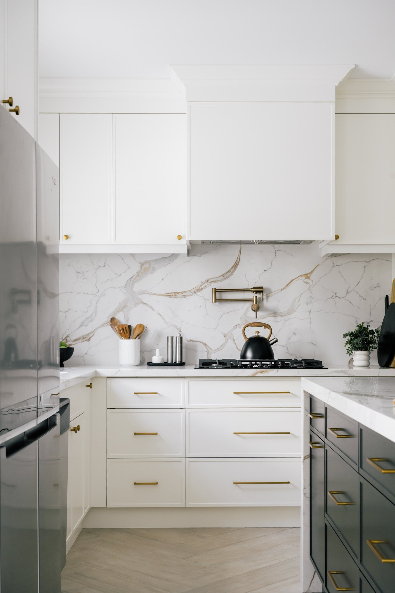 A white kitchen with gold hardware and marbled backsplash. A black kettle sits on the stove.