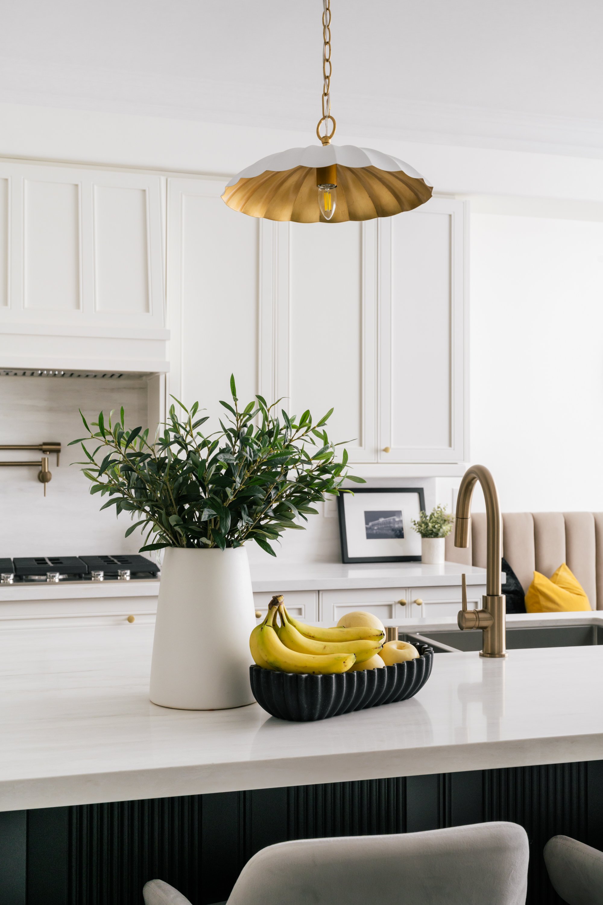 A close up of a vase of greenery and basket of bananas and pears on a kitchen countertop. A white and brass-coloured pendant hangs from above.