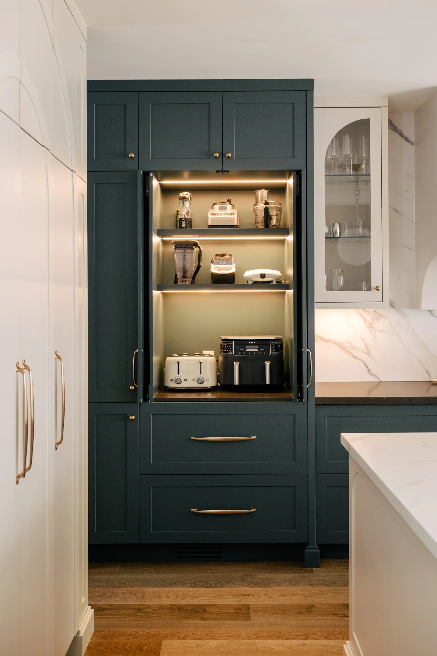 An opened, dark muted turquoise cabinet, displaying countertop appliances neatly stored on 3 shelves with under cabinet lights on.
