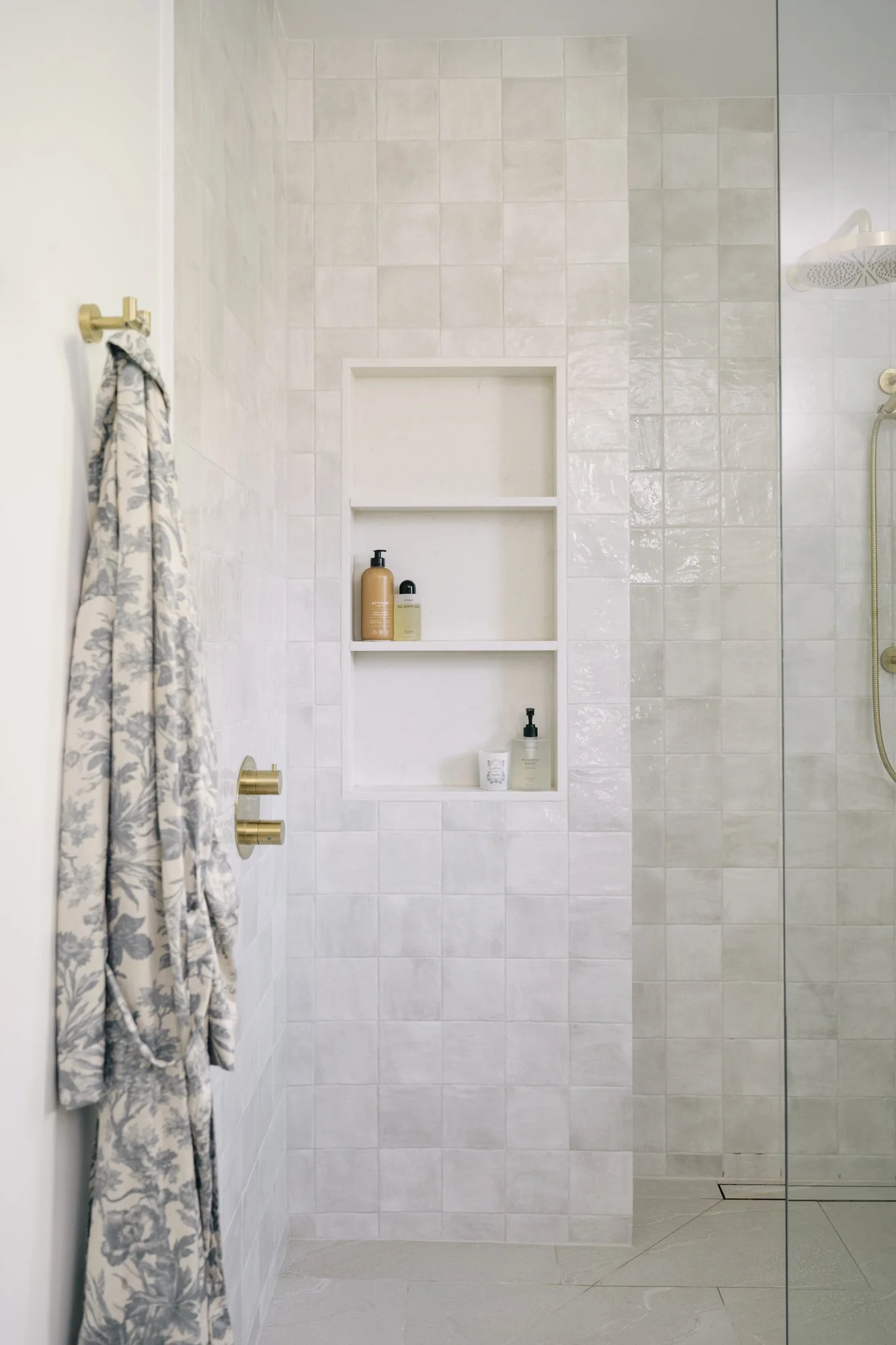 A shower with square tiles and a 3-level wall niche. A floral robe hangs on the wall.