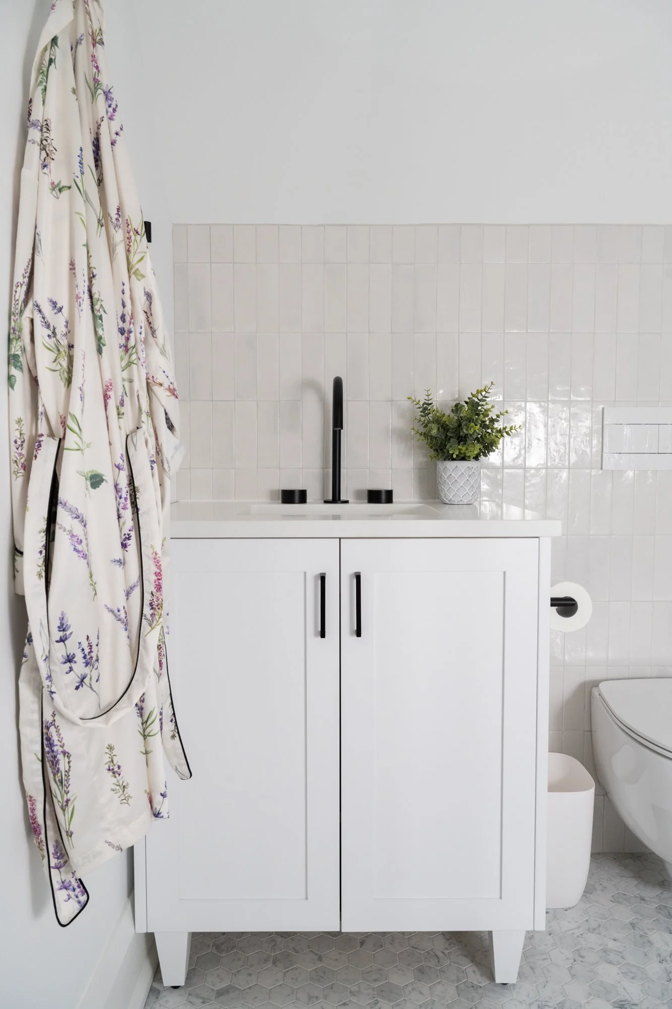 A white bathroom vanity with black hardware. A robe hangs on the wall next to the vanity