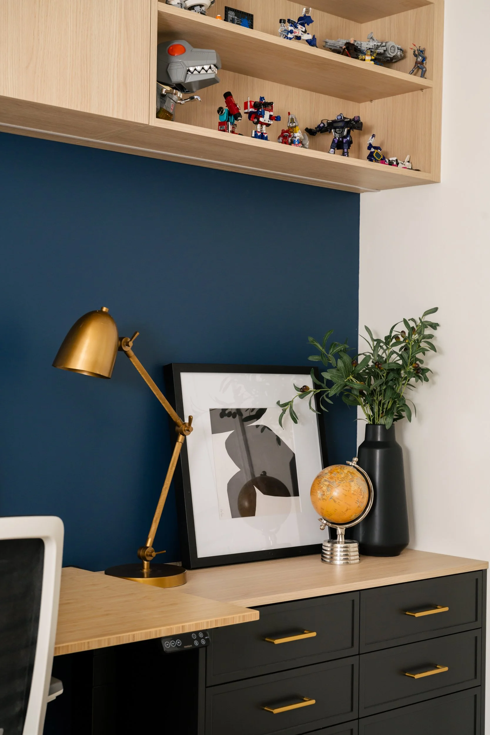 Art frame, lamp, small globe and vase with greenery sits on a shelving unit in a home office