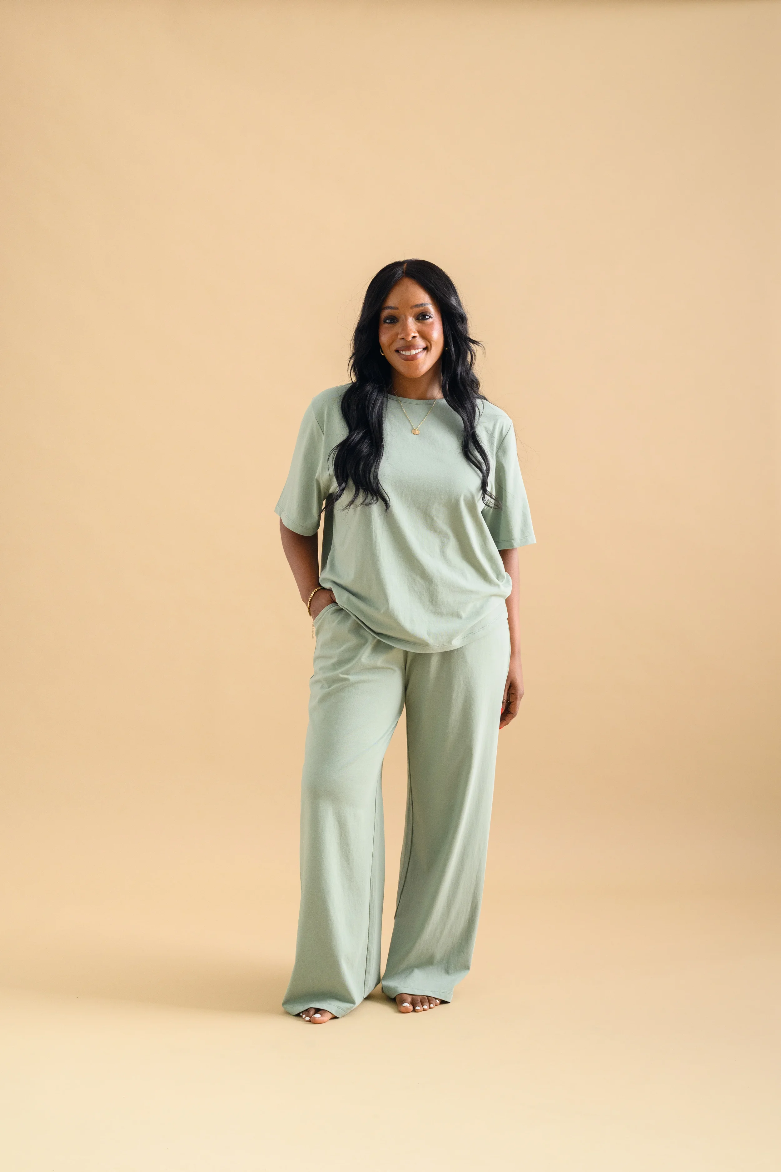 A woman standing against a beige backdrop, wearing a mint green short sleeved shirt and long loose pants by Heirloome House.